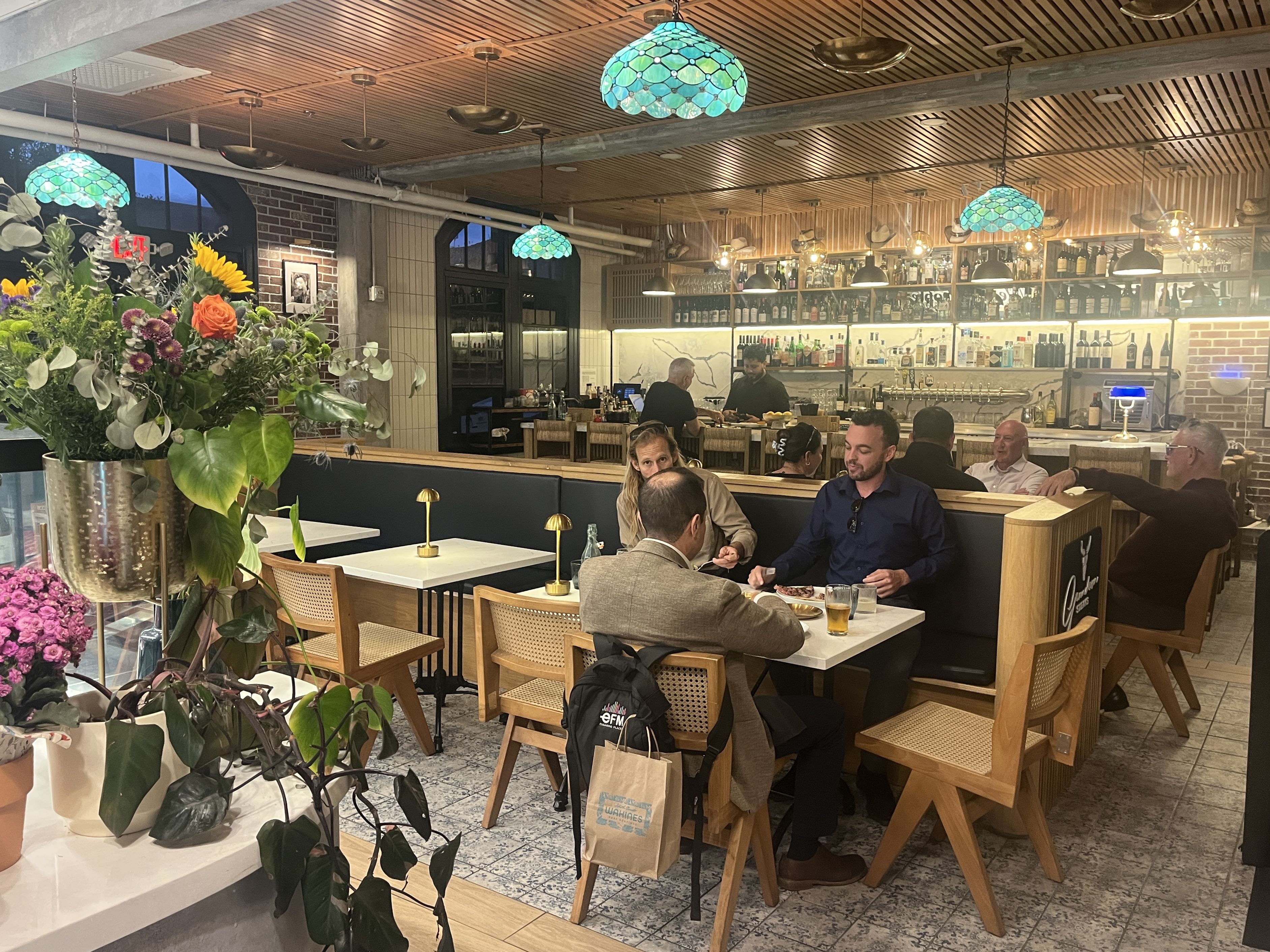 Restaurant interior with blue-green stained-glass pendant lights, a wood slat ceiling, brick accents, and a long bar. Patrons dine at tables; a large floral arrangement sits in the foreground.