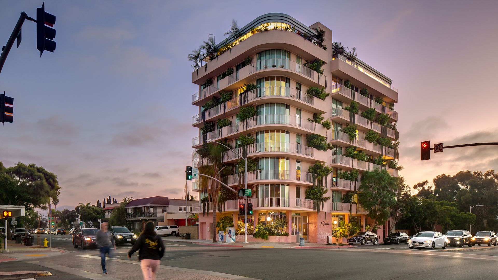 An 8-stroy pink apartment building on the corner of an intersection at sunset.