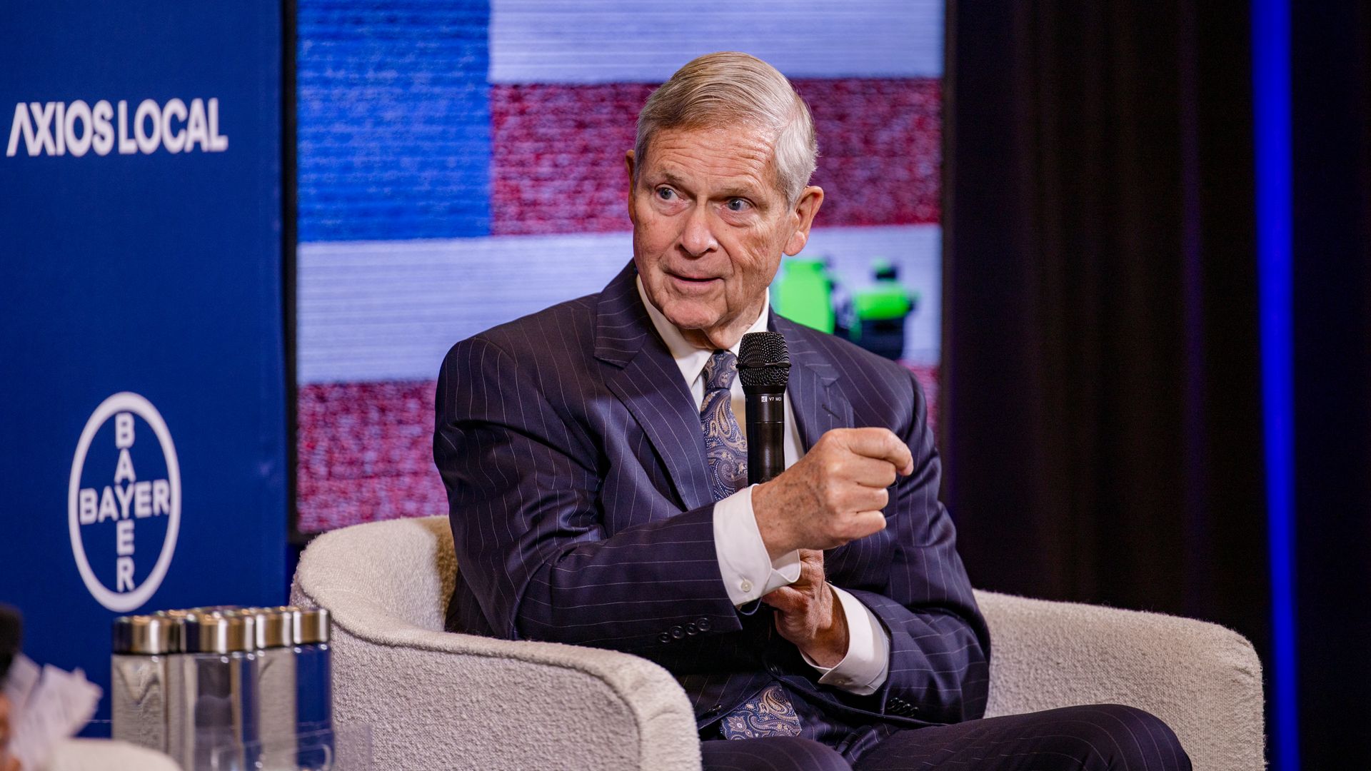An older man in a dark suit sits in a beige chair, holding a microphone on stage, with a blue Axios Local banner and a colorful background behind him.