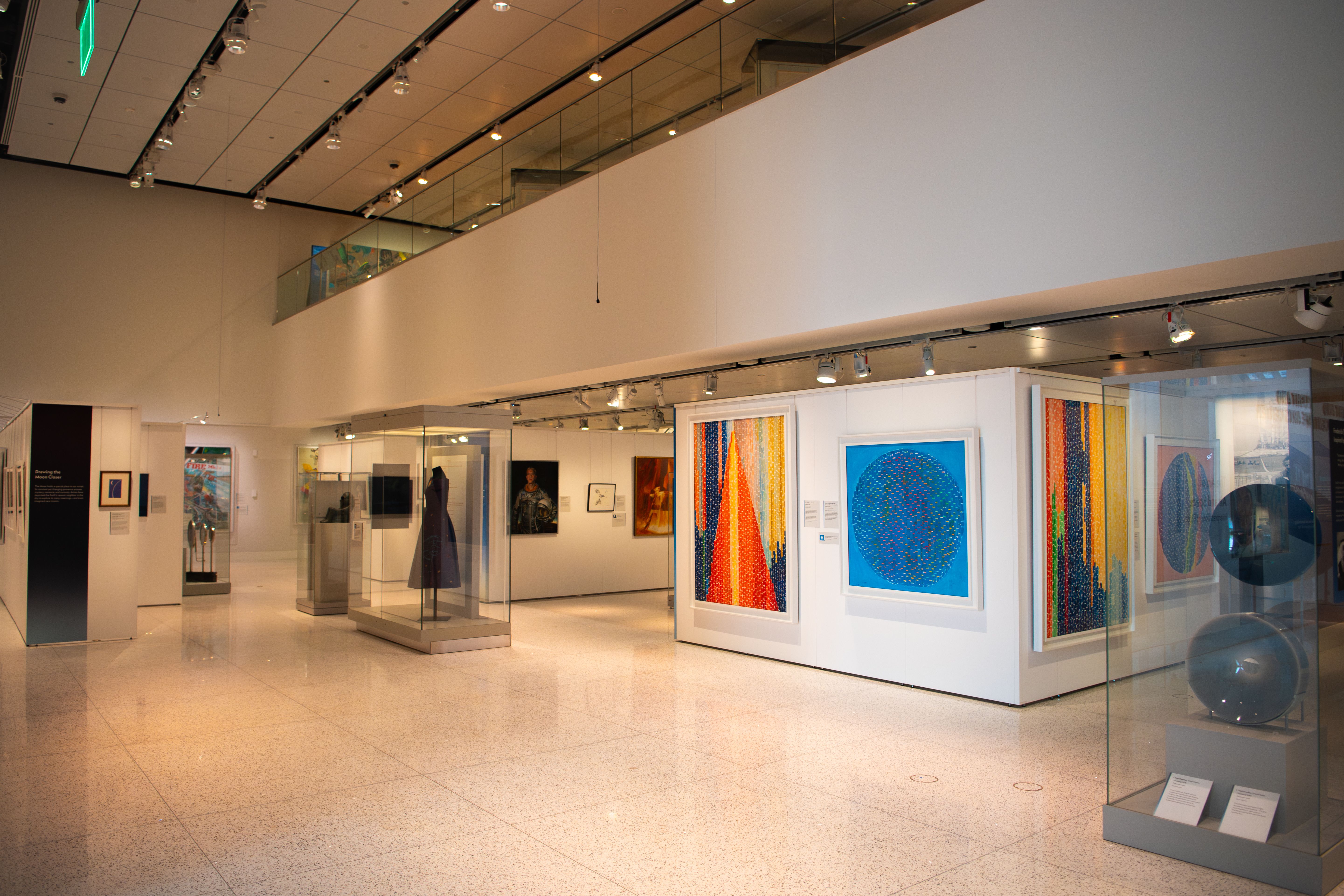 Interior of a modern art gallery: white walls, glass partitions, a glass balcony, polished tiled floor, and bright colorful framed artworks along the walls under track lighting.