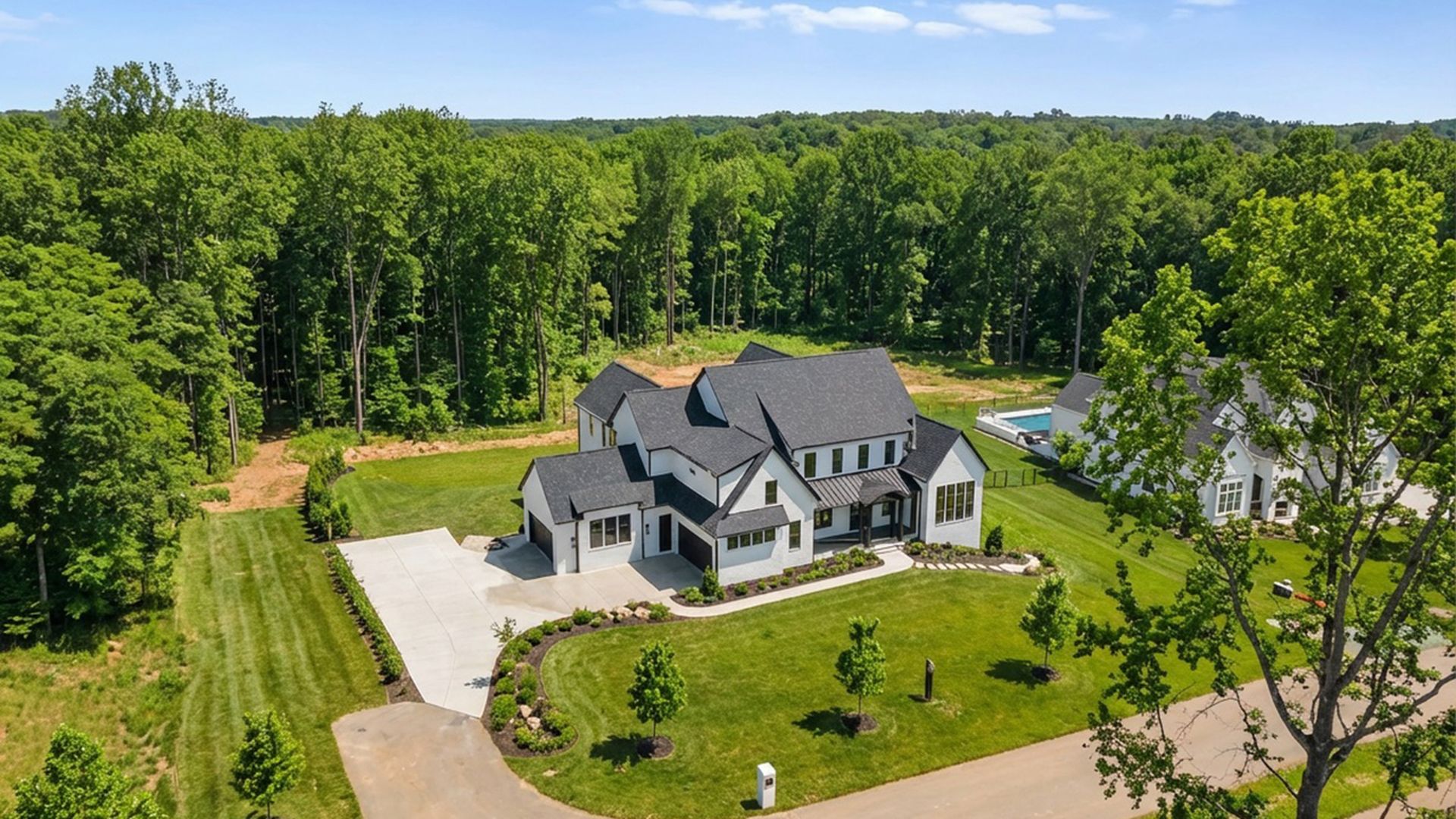 A large modern white house with black roof, surrounded by a green lawn, young trees, and a forest in the background, under a clear blue sky.