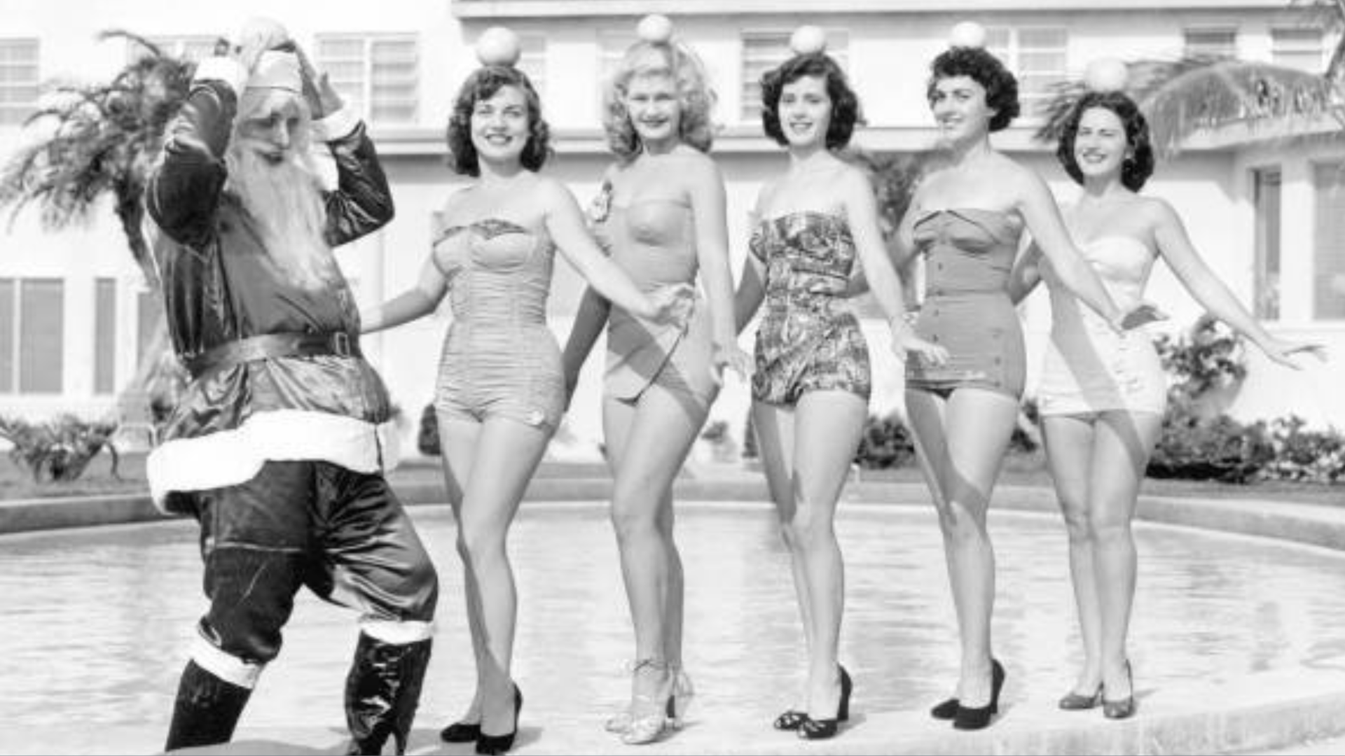 A black-and-white photo of a man dressed as Santa Claus and five women in vintage bathing suits posing on the edge of a pool.