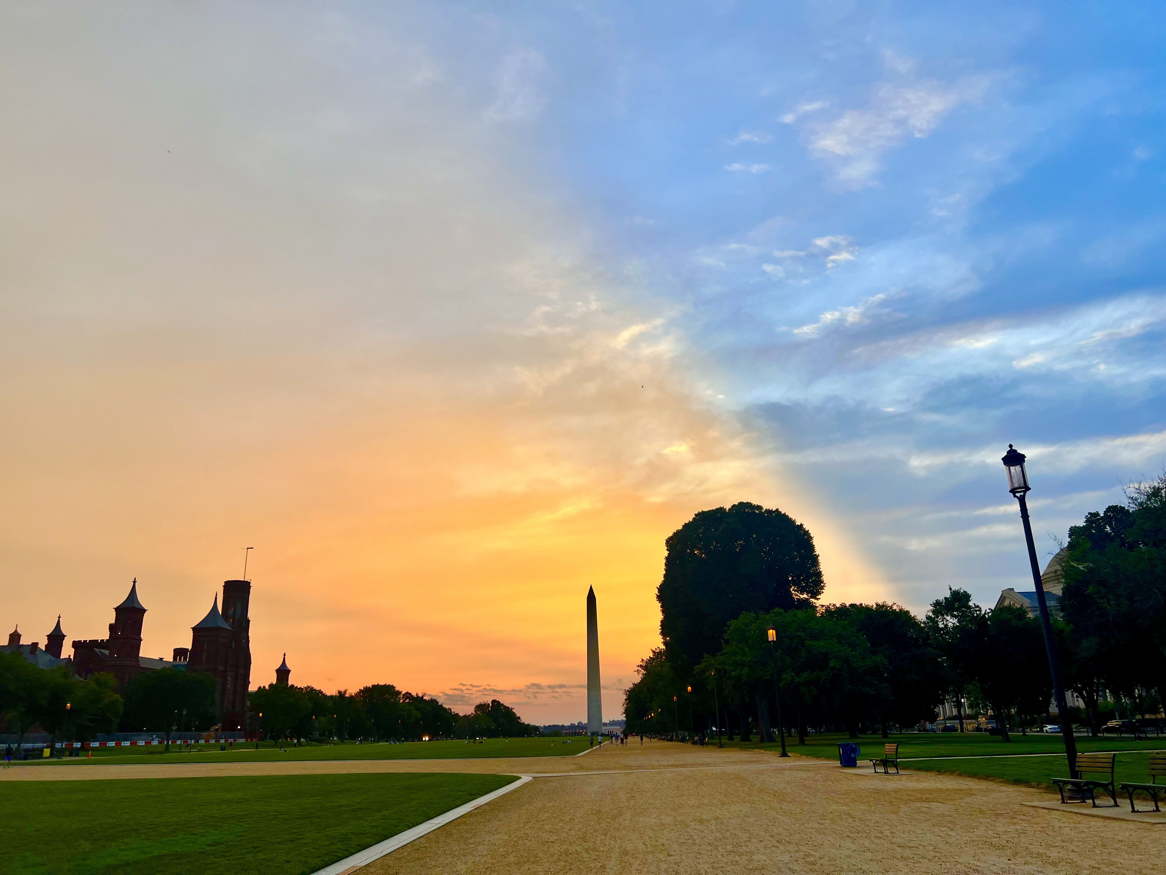 Sunset over the Washington Monument with orange and blue sky, green grass, trees, lampposts, and benches along a wide path in a park setting.