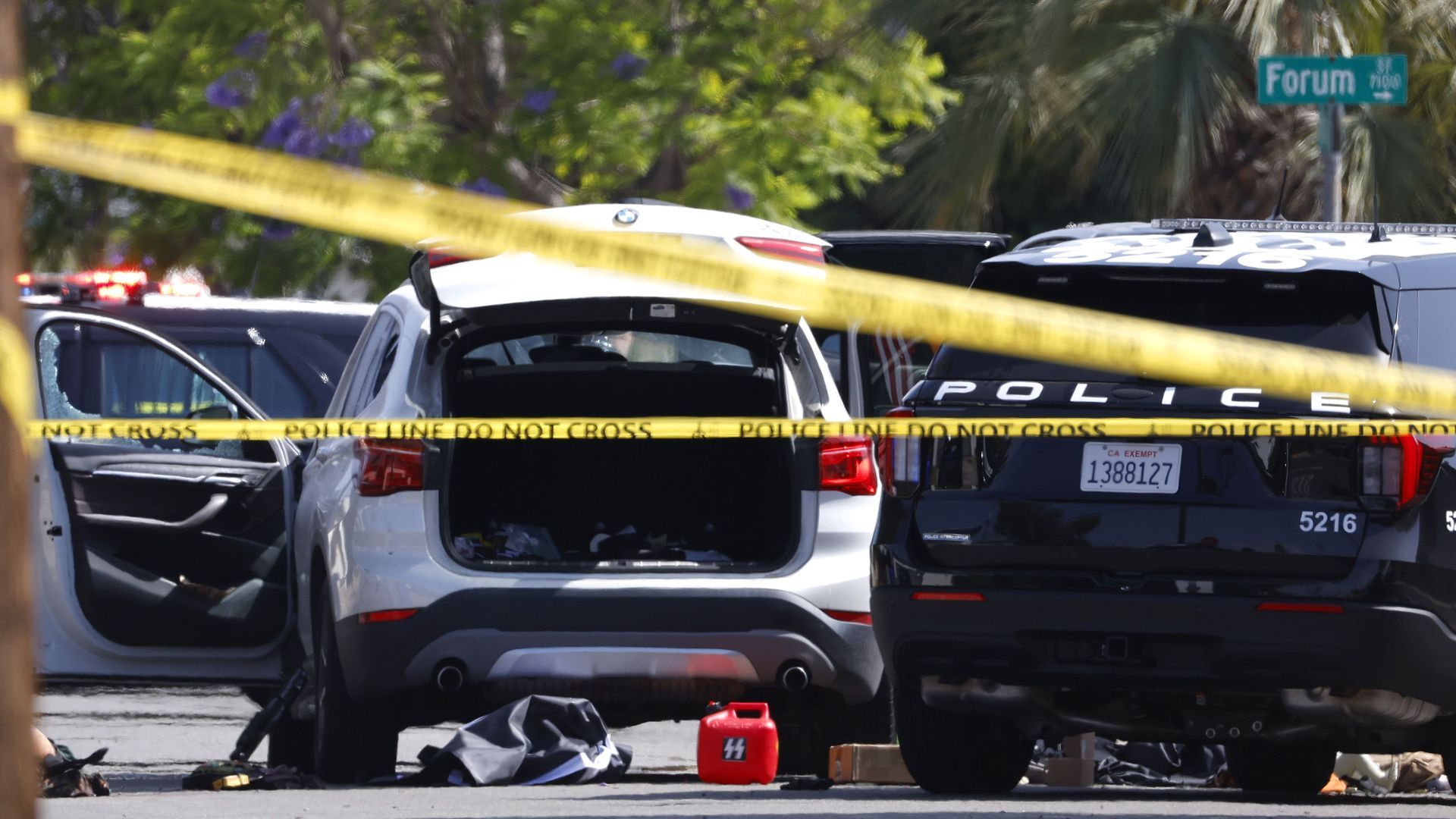 Police tape reads "POLICE LINE DO NOT CROSS" across the foreground. A white SUV with its trunk open and BMW emblem visible with a red gas can on the ground and firearm leaning against the driver's seat; police cars parked nearby amid green trees and purple flowers.