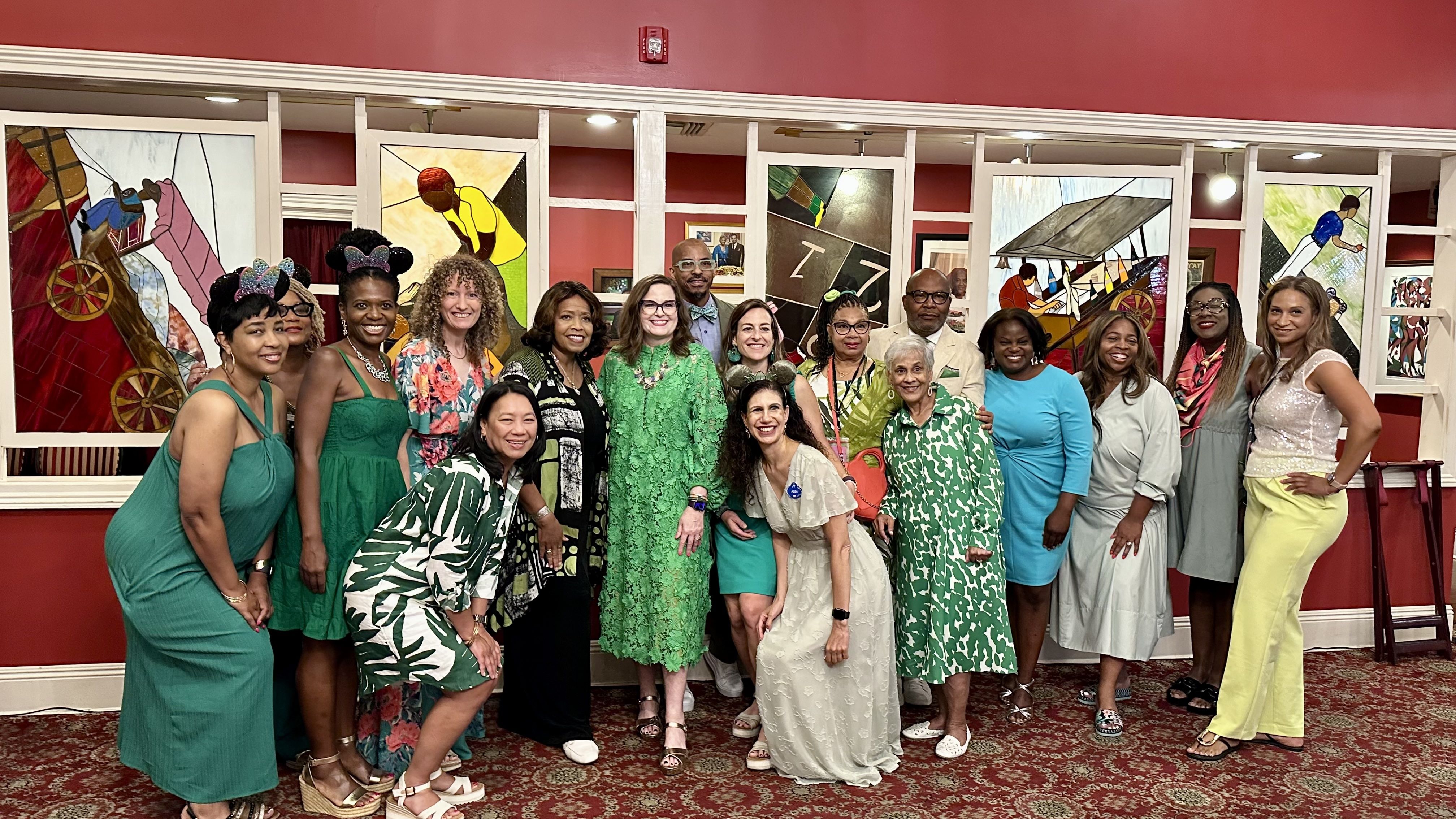 Group photo of sixteen people, mostly women, dressed in shades of green, blue, yellow, and white, standing and posing indoors in front of stained glass panels with colorful artistic designs.