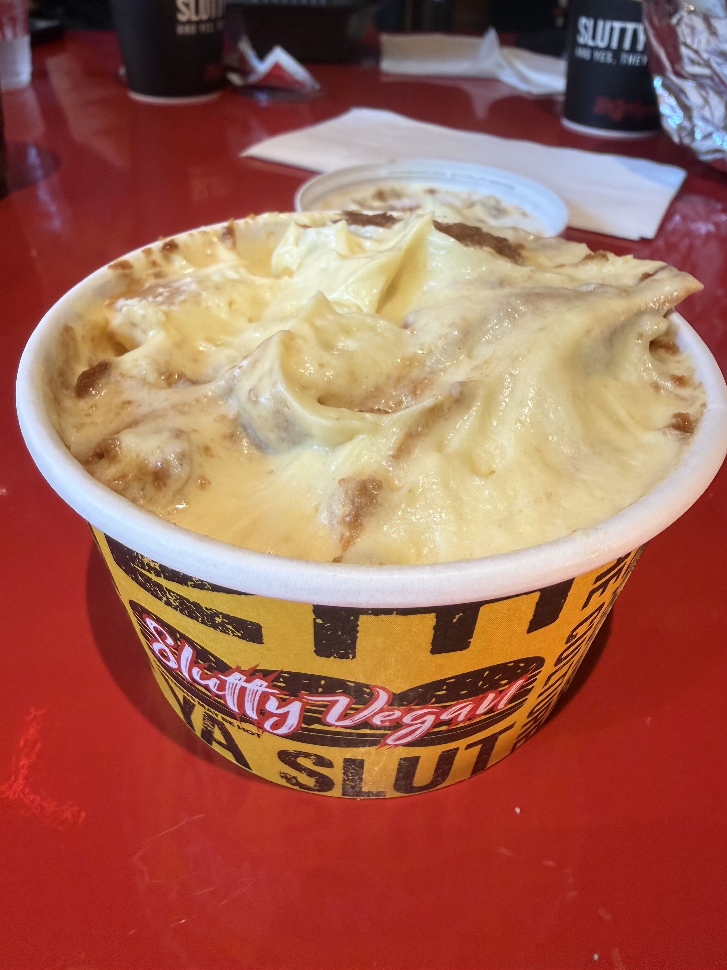 Close-up of a large yellow Slutty Vegan cup on a red table, overflowing with a creamy vanilla dessert topped with whipped cream; blurred napkins and other cups visible in the background.