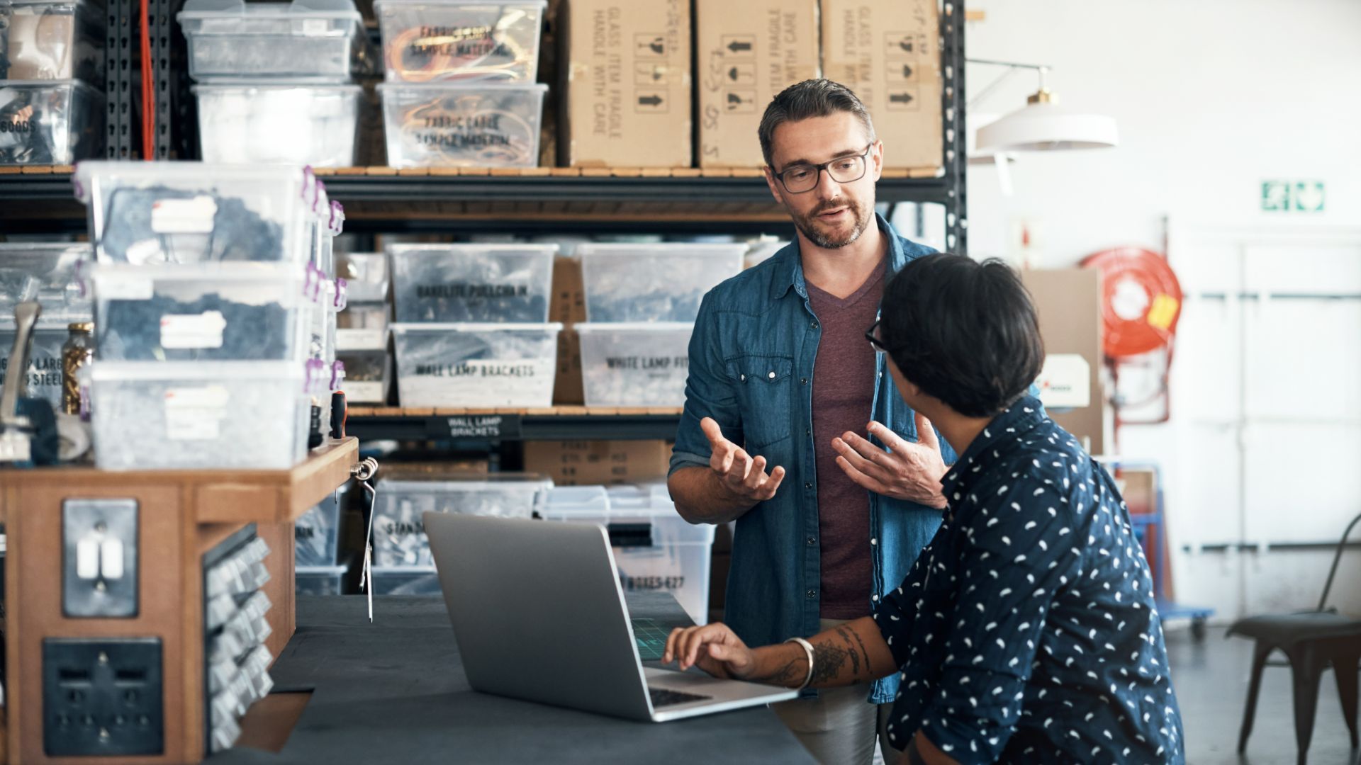 Two colleagues discuss work in a supply room, with storage bins and boxes in the background and a laptop open on the counter.