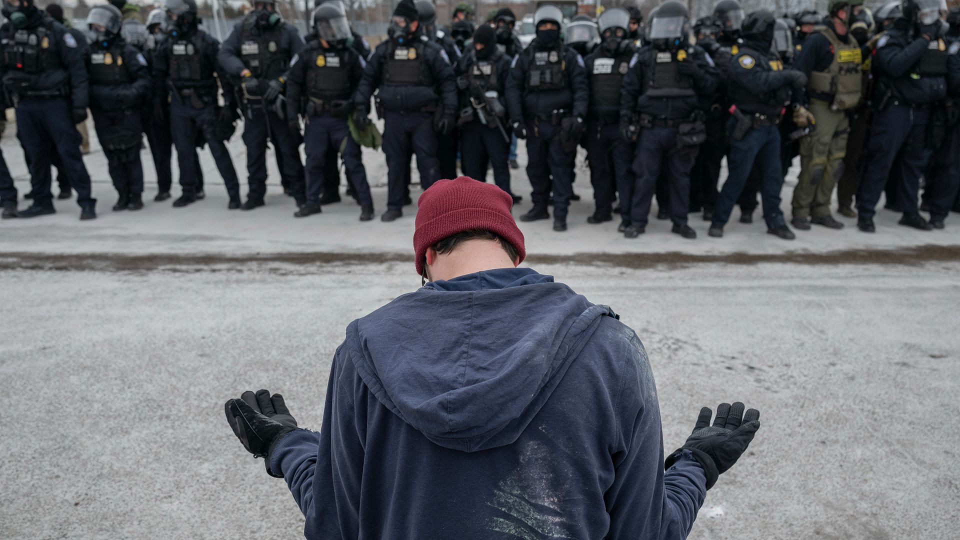 a man kneels in front of a line of ICE officers in tactical gears