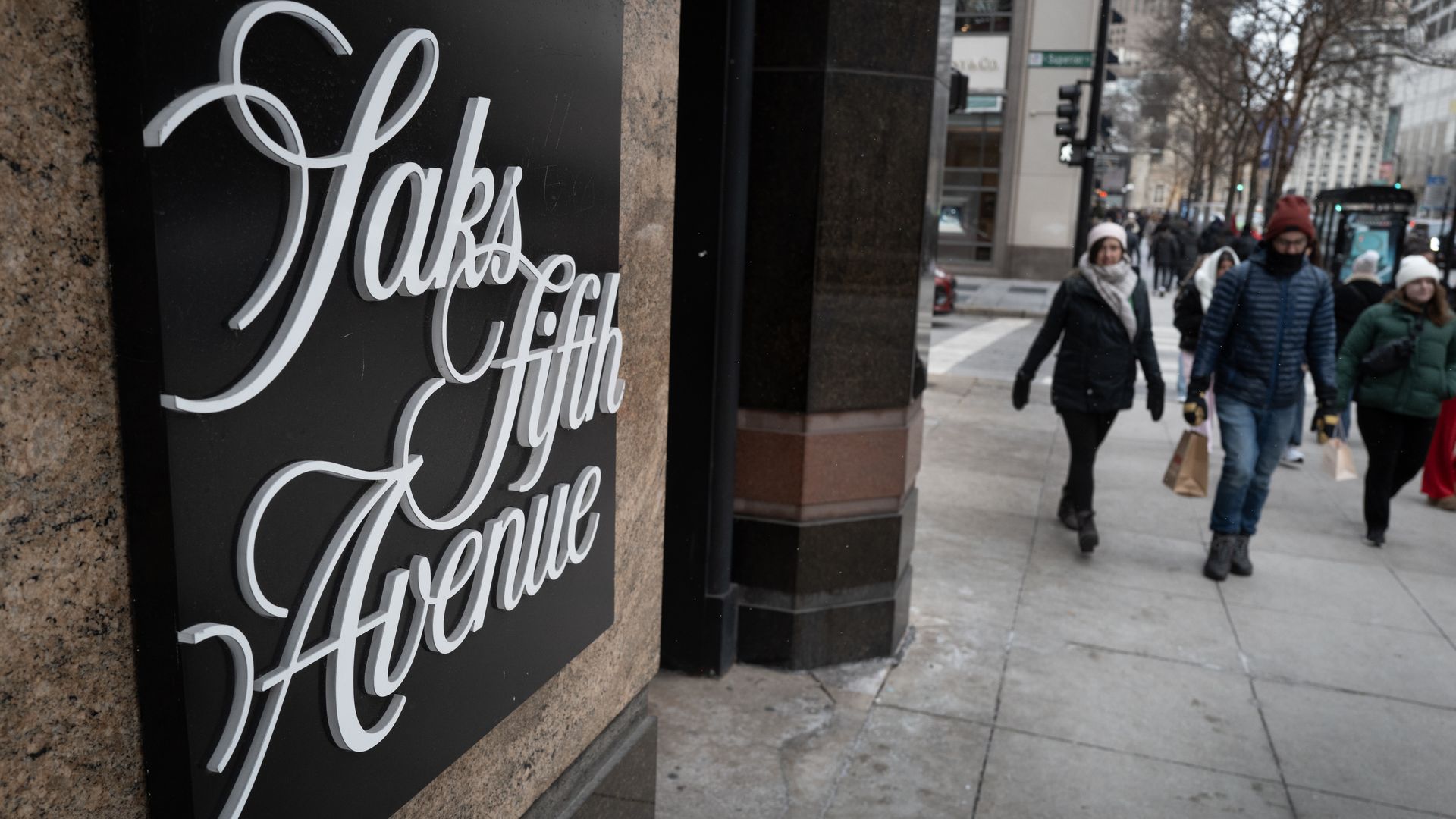 Close-up of the black "Saks Fifth Avenue" sign on a stone building, as a winter city street scene unfolds with pedestrians in coats walking along the sidewalk.