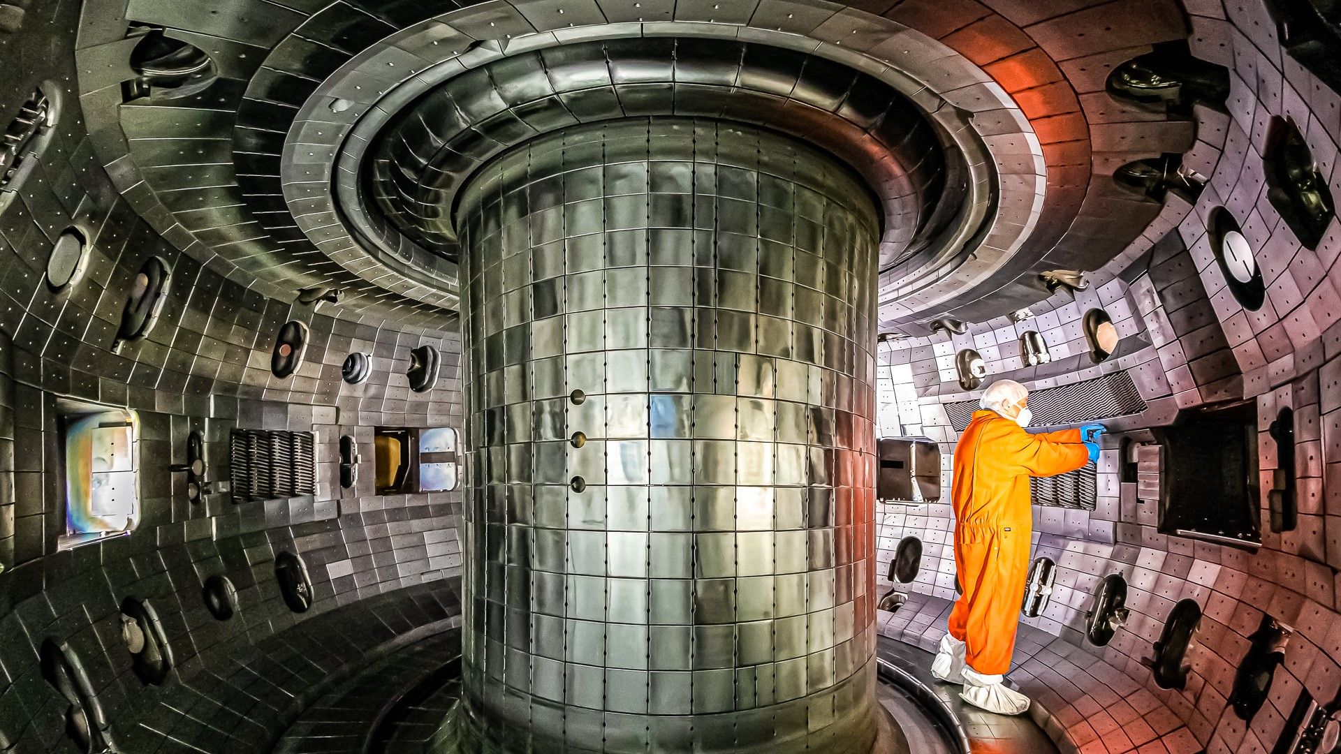 Inside a large metallic circular chamber with a central cylindrical column; a worker in an orange protective suit and white helmet stands to the right, inspecting equipment.