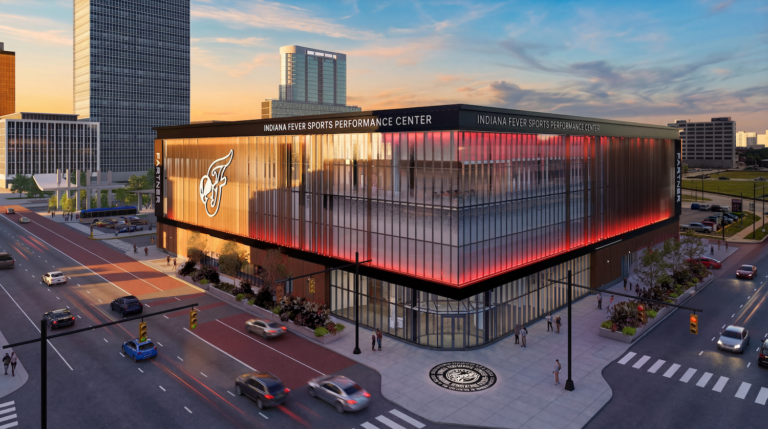 Modern glass building labeled "Indiana Fever Sports Performance Center" at a busy city intersection at sunset, illuminated by red neon along the edges; Fever logo on the side with pedestrians and moving cars nearby.