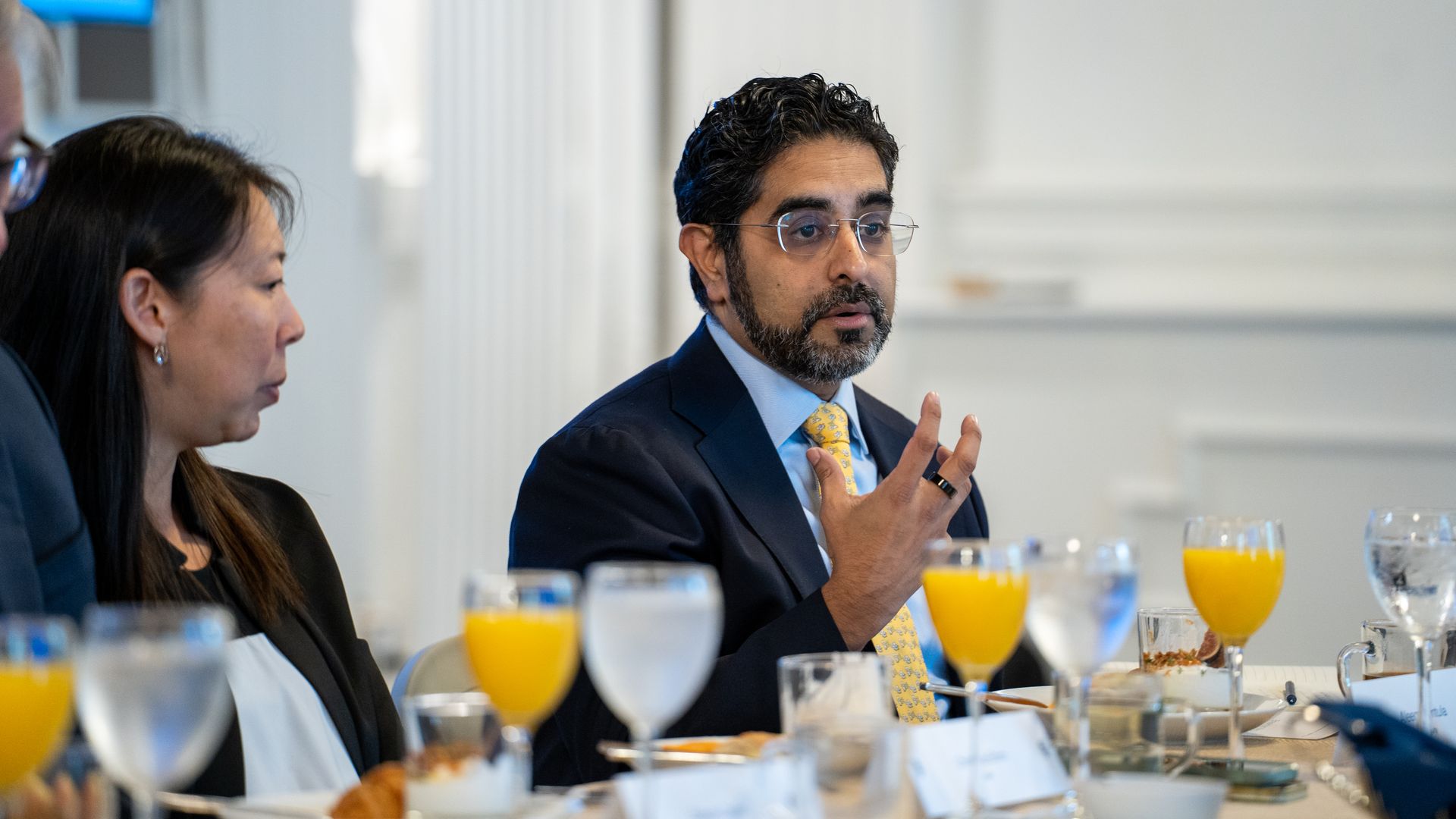 Bearded man in a dark suit and yellow tie, wearing glasses, speaks with hand gestures at a breakfast meeting; orange juice glasses, water, and plates visible on the table.