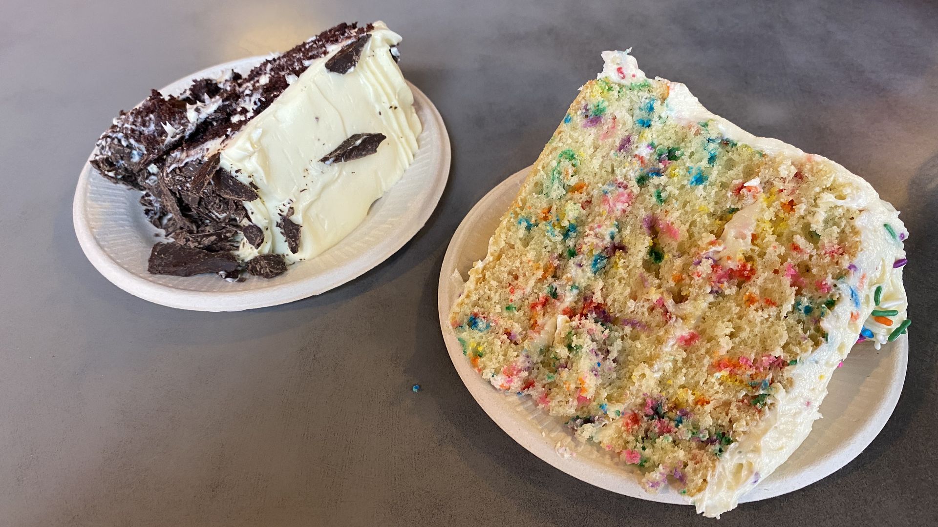 A slice of chocolate cake with white frosting on a paper plate at left, at right a slice of vanilla cake with rainbow sprinkles baked in, plus white frosting with sprinkles