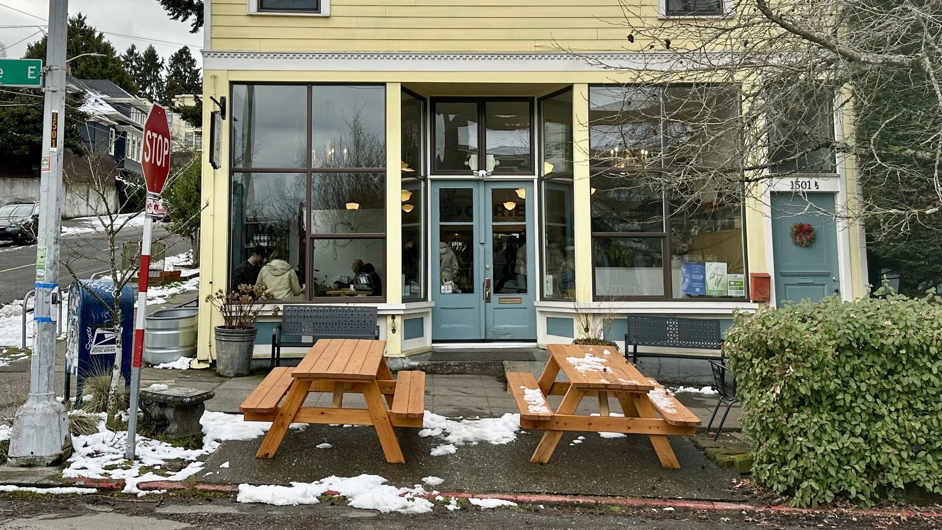 Yellow building with teal double doors and large windows showing people inside. Two wooden picnic tables with patches of snow on a wet sidewalk. Stop sign and street sign for 15th Ave E.