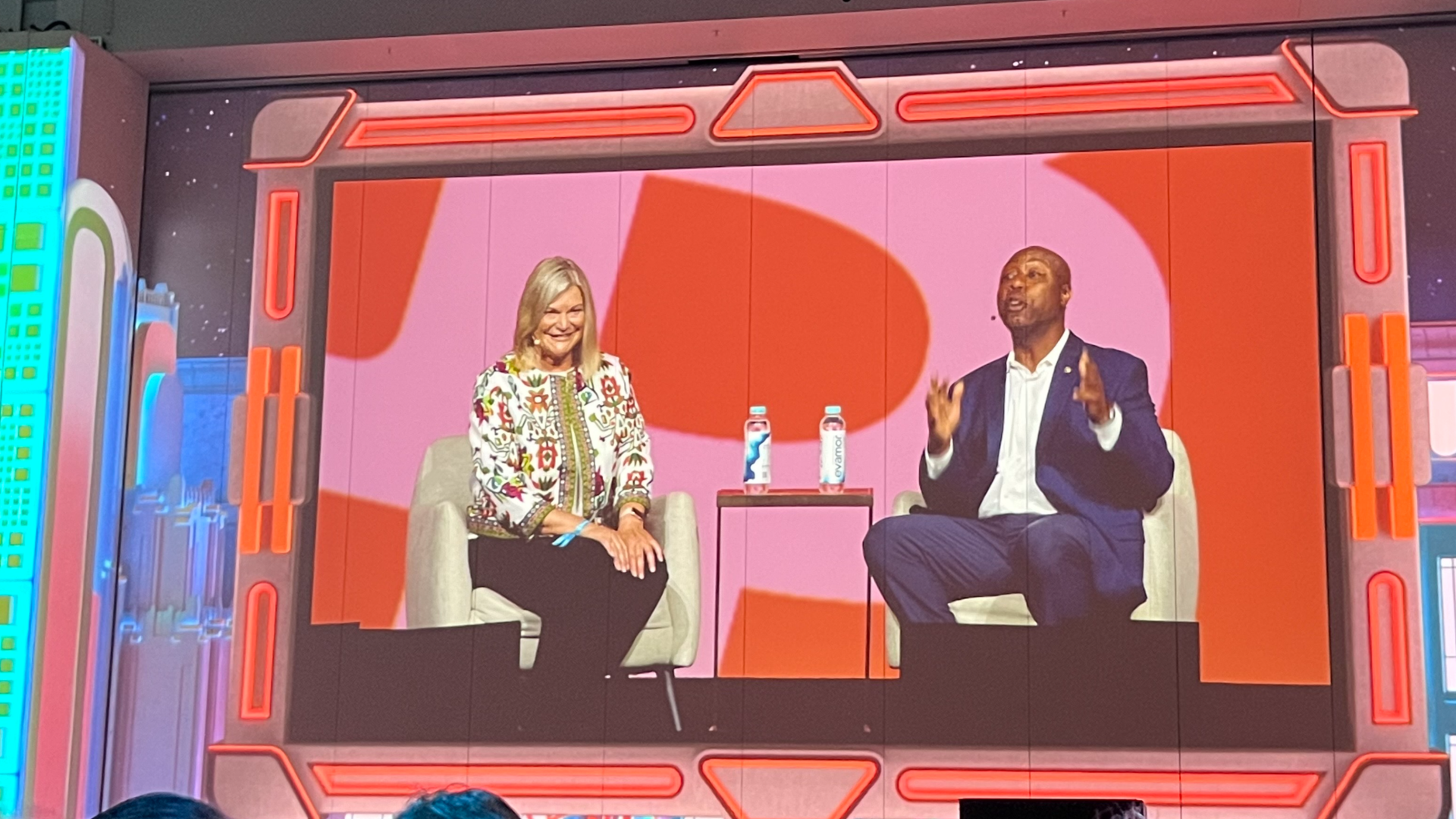 Two people speaking at a conference, a man and woman, shown on a massive screen.