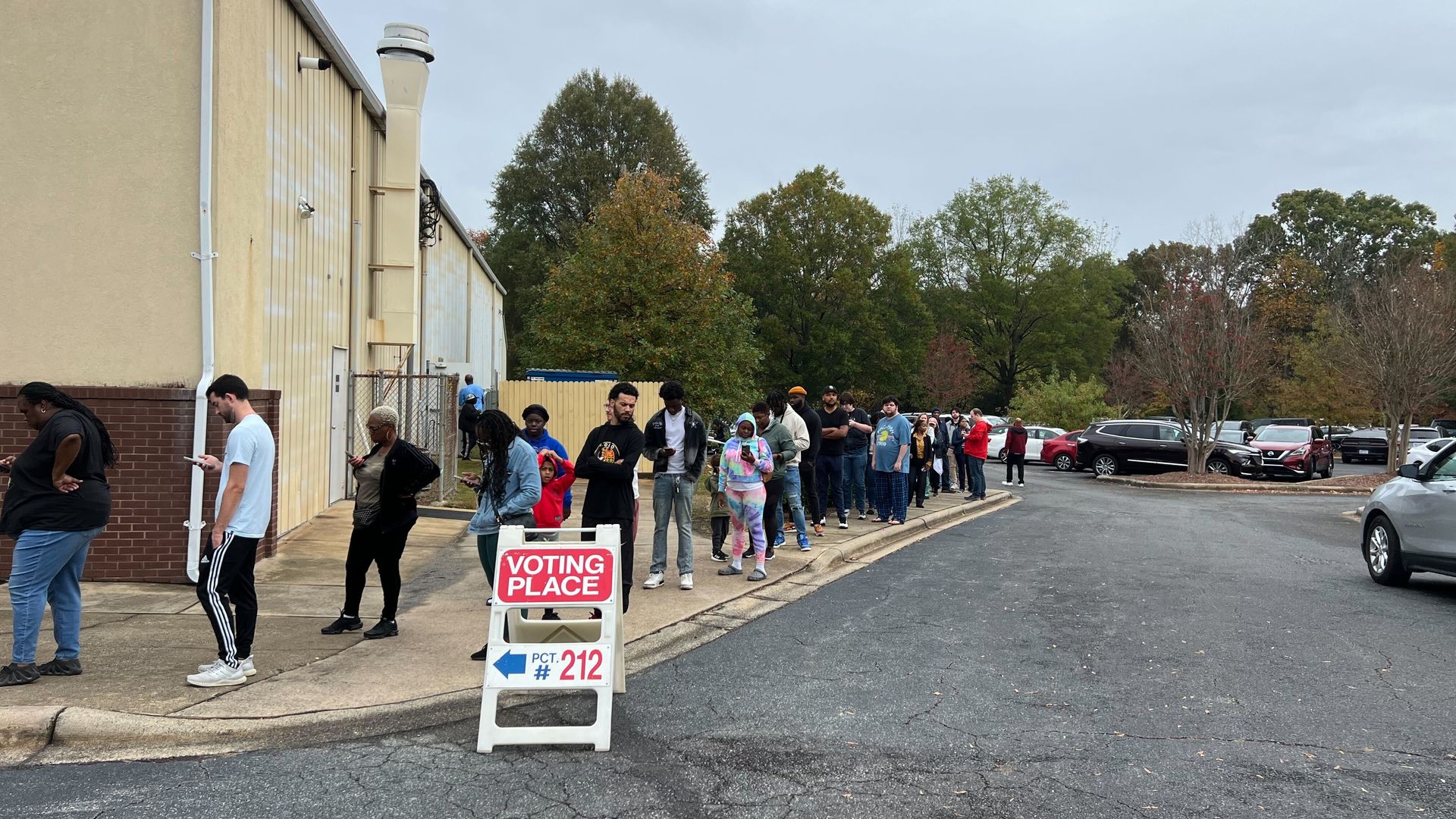 people in line. a sign that reads "voting place" stands near them