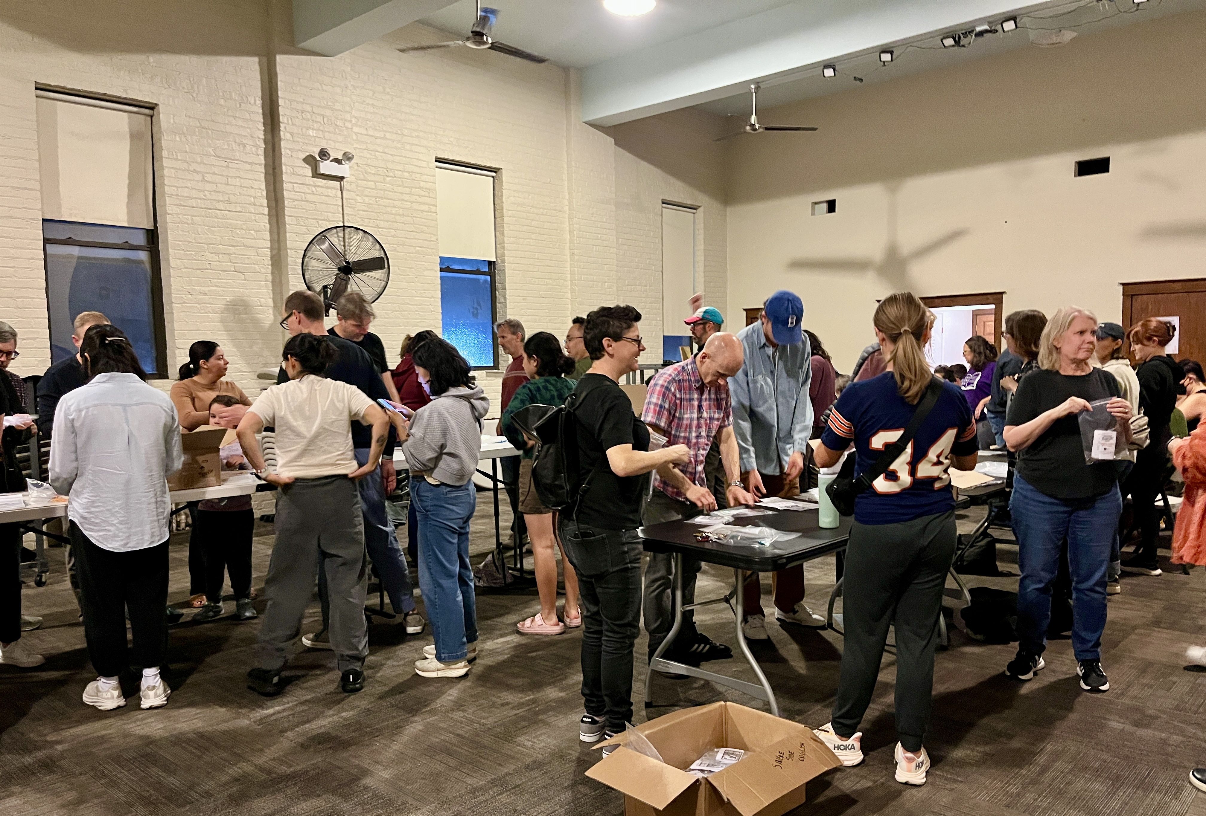 A diverse group of people standing and interacting in a large room with white brick walls, folding tables, and a large fan on the wall, possibly organizing or distributing items.