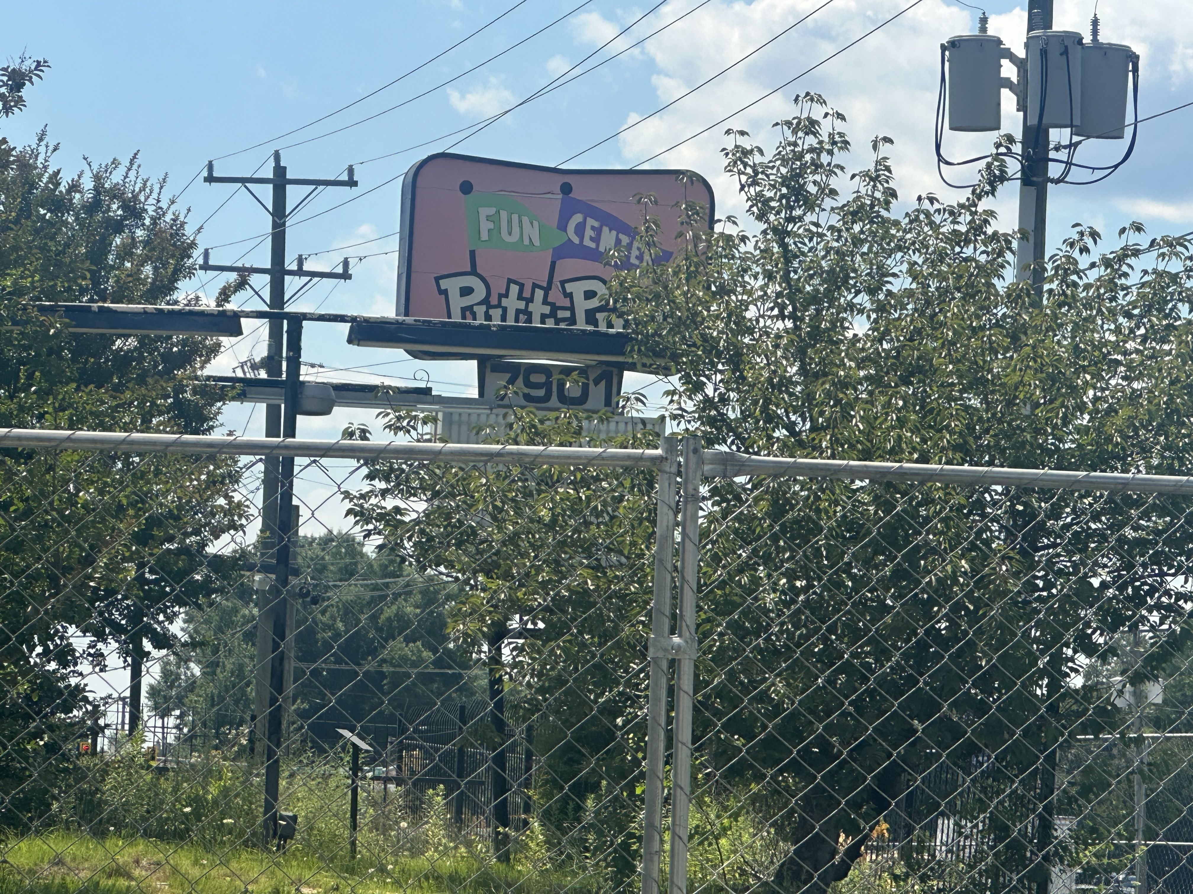 Pink sign reading FUN CENTER and Putt-Putt partially visible behind a chain-link fence, with power lines and transformers on a pole, and leafy trees against a blue sky.