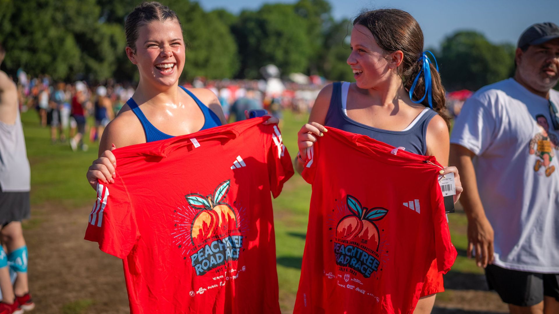 Two smiling young women in athletic wear holding red Peachtree Road Race shirts outdoors on a sunny day with a crowd and trees in the background.