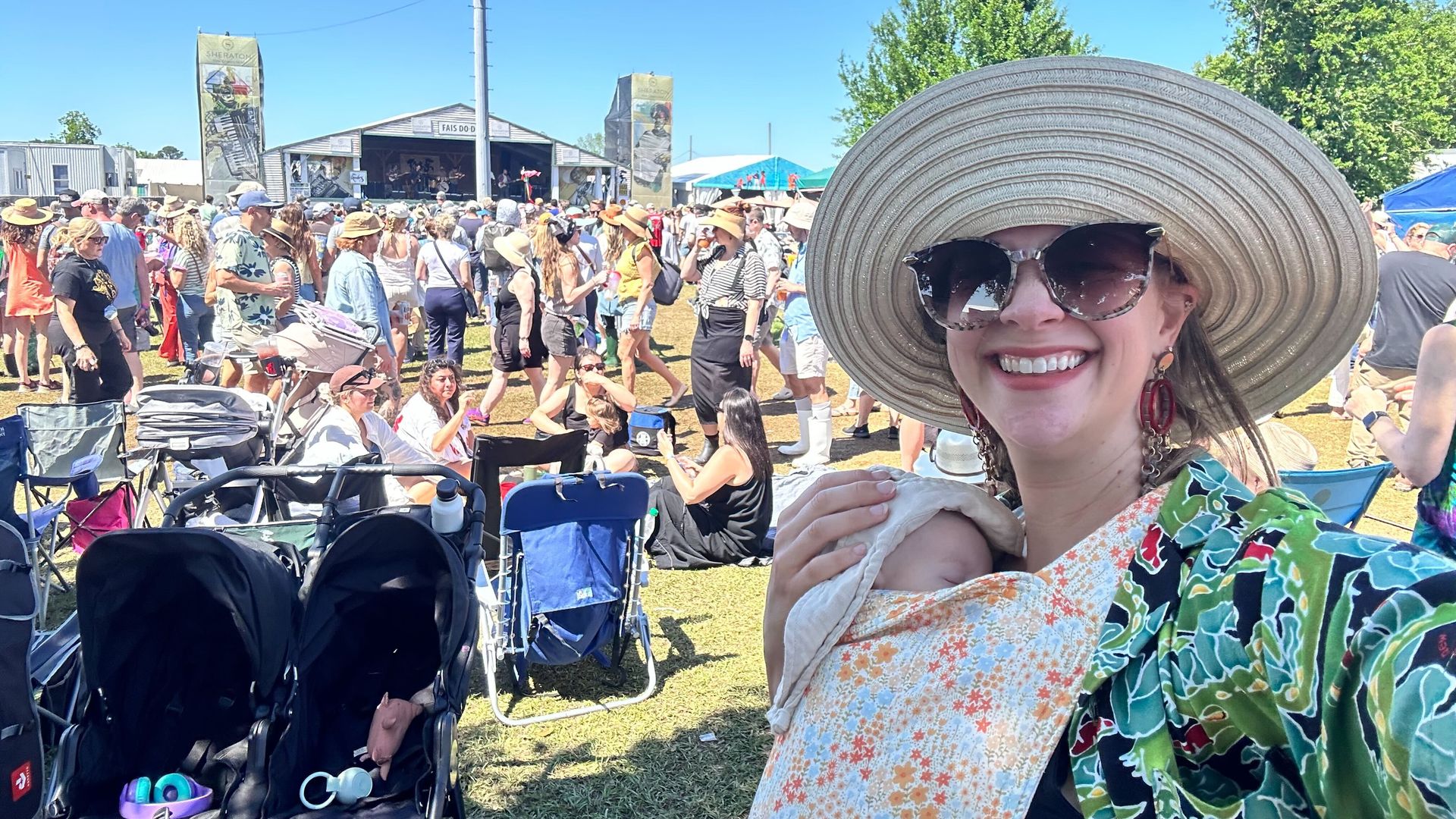 Crowd at a sunny outdoor festival under a blue sky, stage and banners in the background; a smiling person in the foreground wears a wide-brimmed hat and sunglasses.