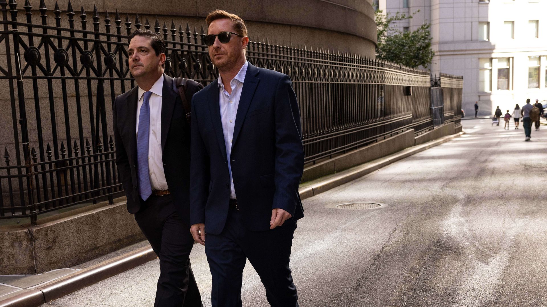 Two men walking on a small street along a gothic fence in business suits, heading from light to shadow.