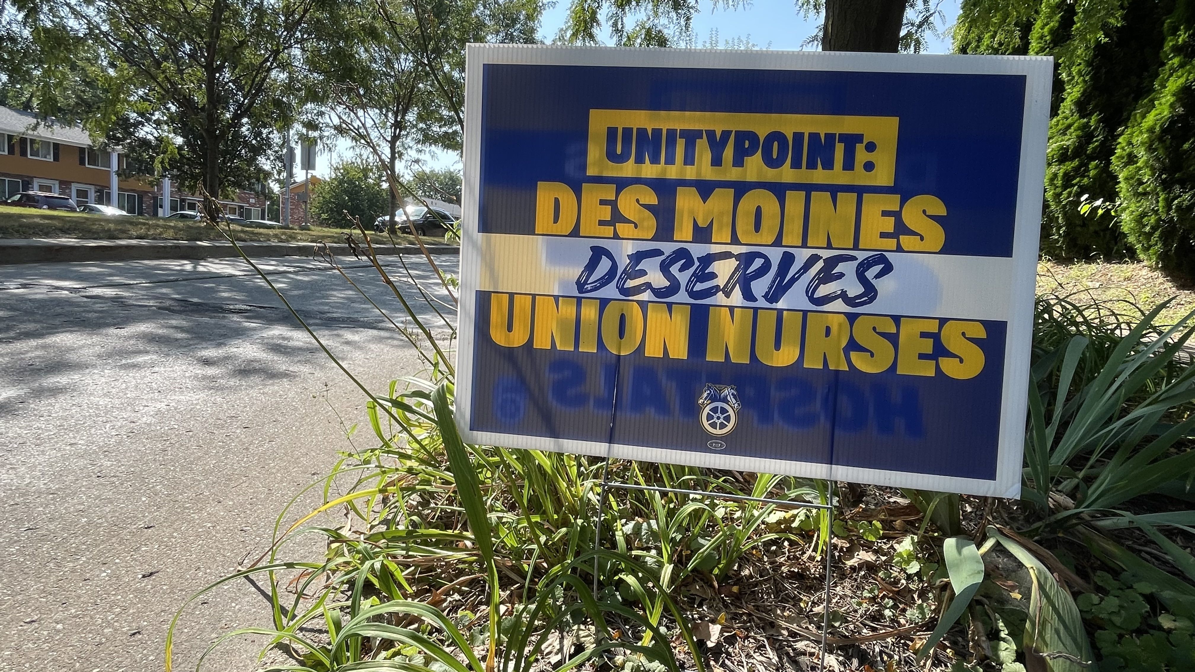 Yard sign in grass with blue and yellow text reading "UNITYPOINT: DES MOINES DESERVES UNION NURSES" in a sunny suburban area with trees and houses nearby.