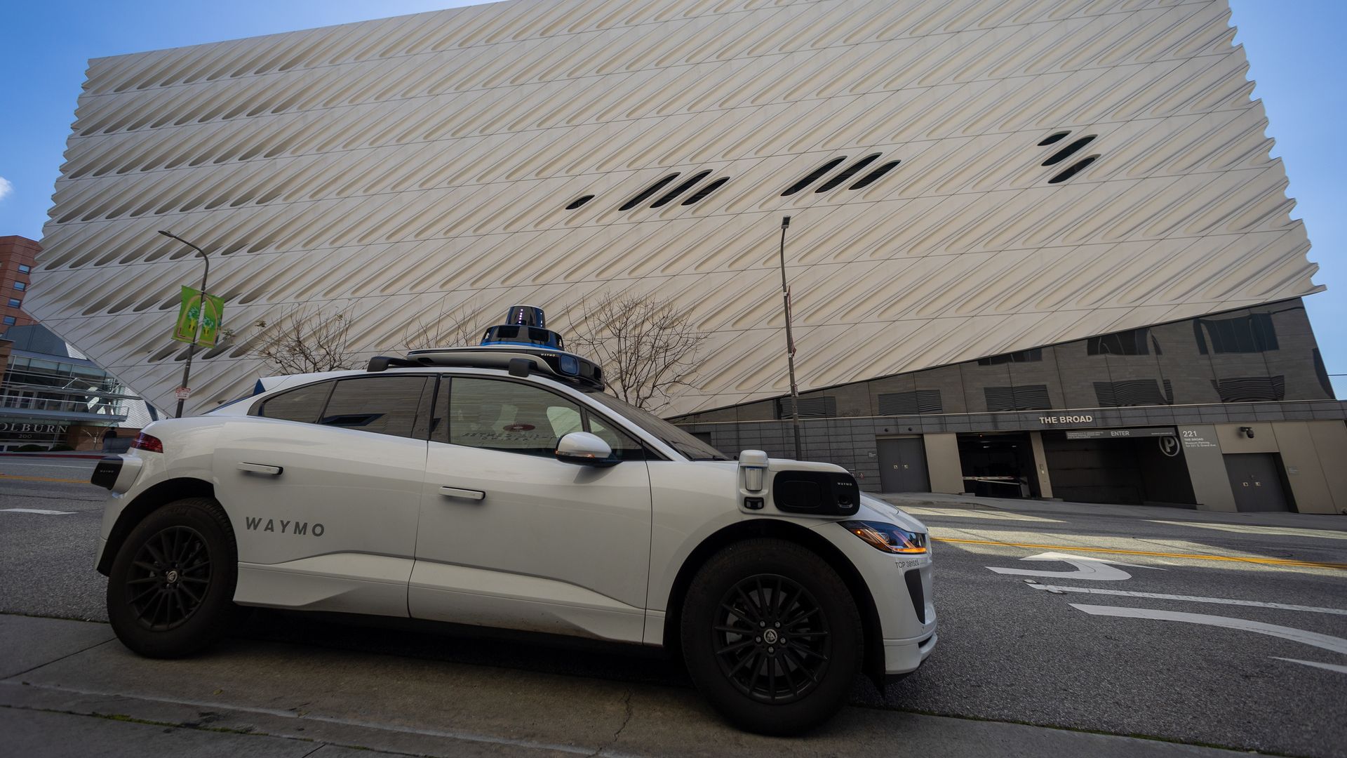 A white sedan outfitted with cameras drives past a Brutalist-inspired building.
