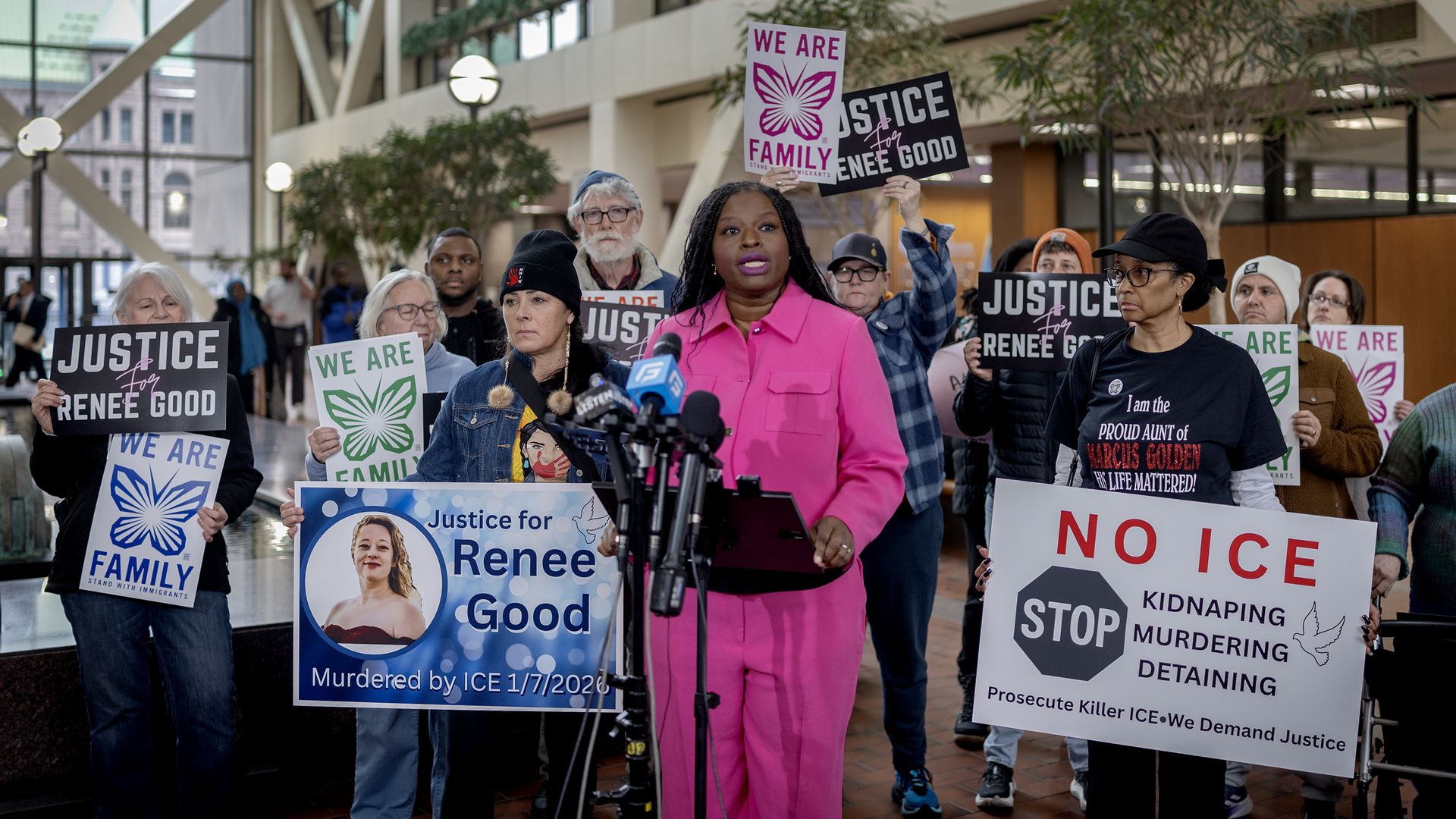 Nekima Levy Armstrong stands in a pink outfit at microphones, behind her are people holding signs against ICE and asking for justice for Renee Good. 