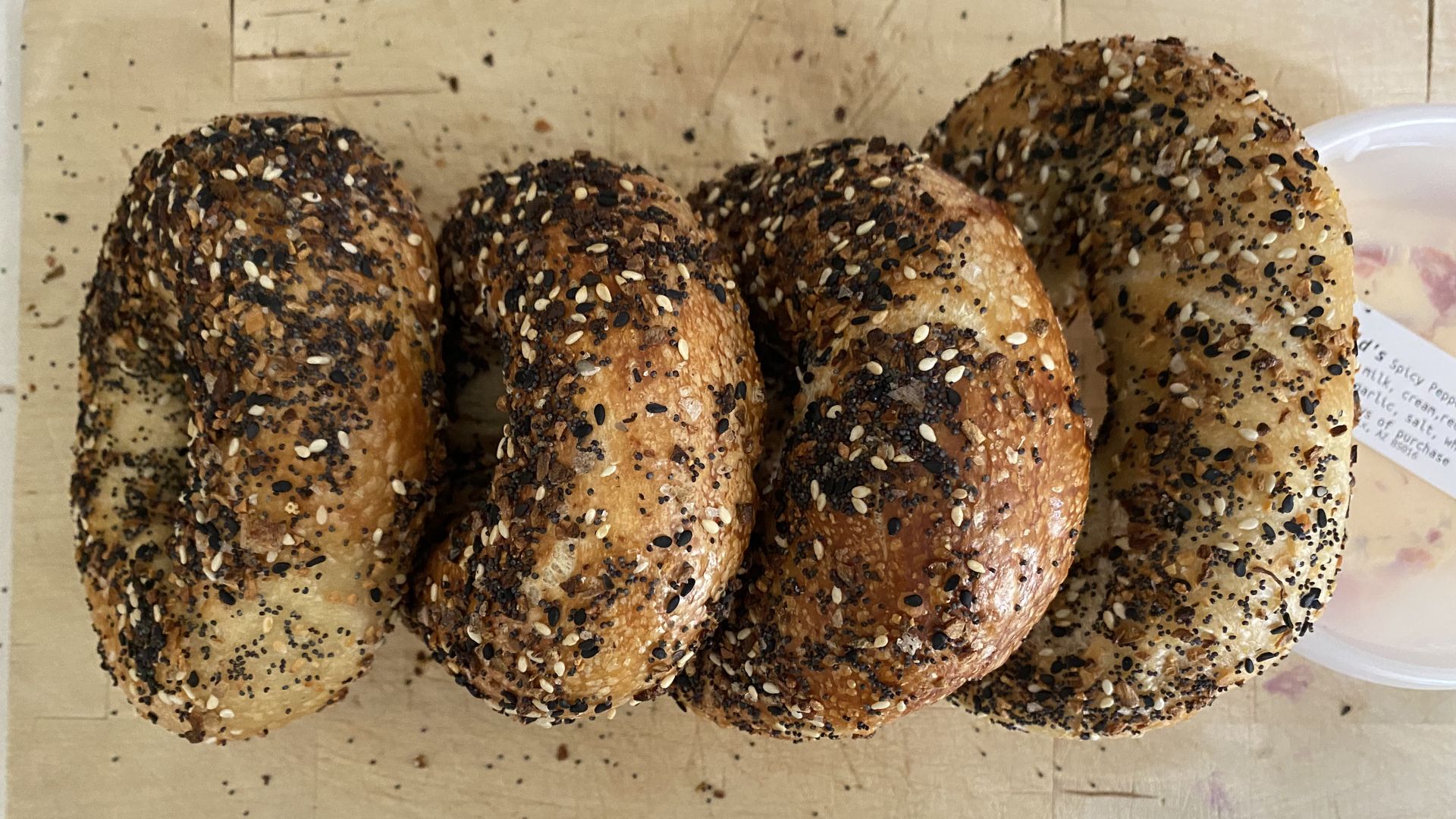 Four everything bagels in a row on a wooden cutting board next to a plastic container of cream cheese.