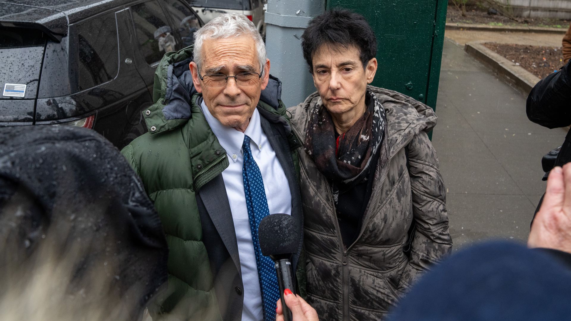 Photo of an older man and woman, standing outside in winter coats, looking pained with a microphone directed at them.