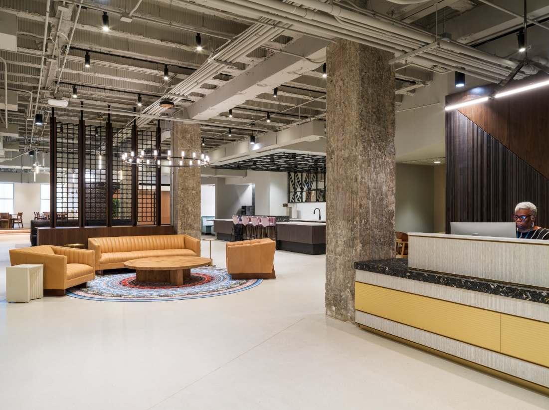 Modern office lobby with tan leather couches around a colorful round rug, concrete pillars, open ceiling with visible pipes, and a reception desk with a person working on a computer.