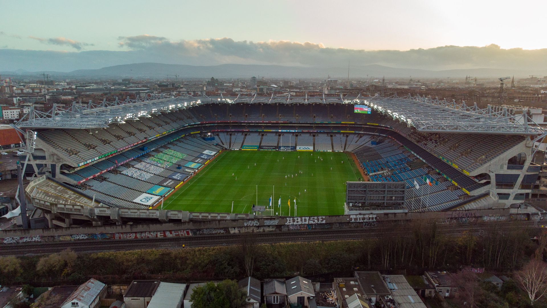 An aerial photo showing a soccer stadium with green grass, seats, and the city sprawling in the background 