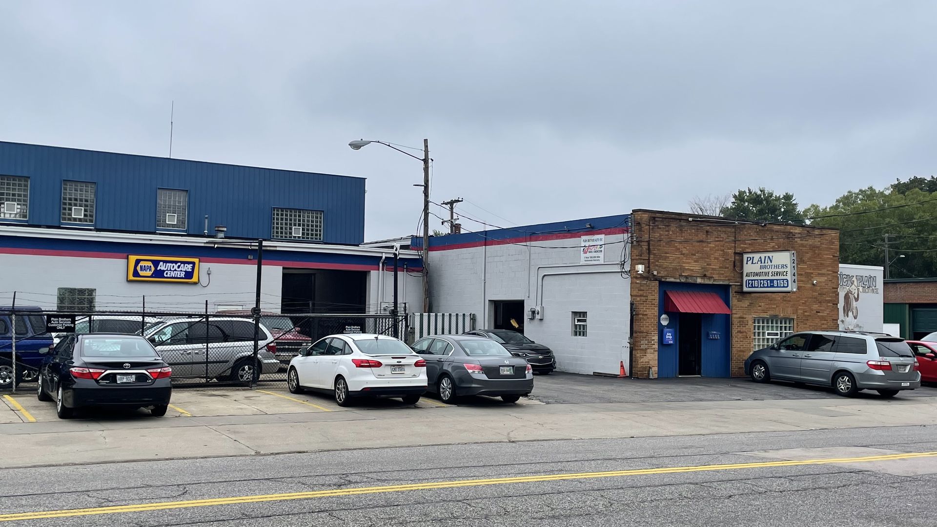 A street view of auto shops: a blue building with a NAPA AutoCare Center sign, a brick shop on the right, and several cars parked in front. cloudy sky and utility poles in the scene.