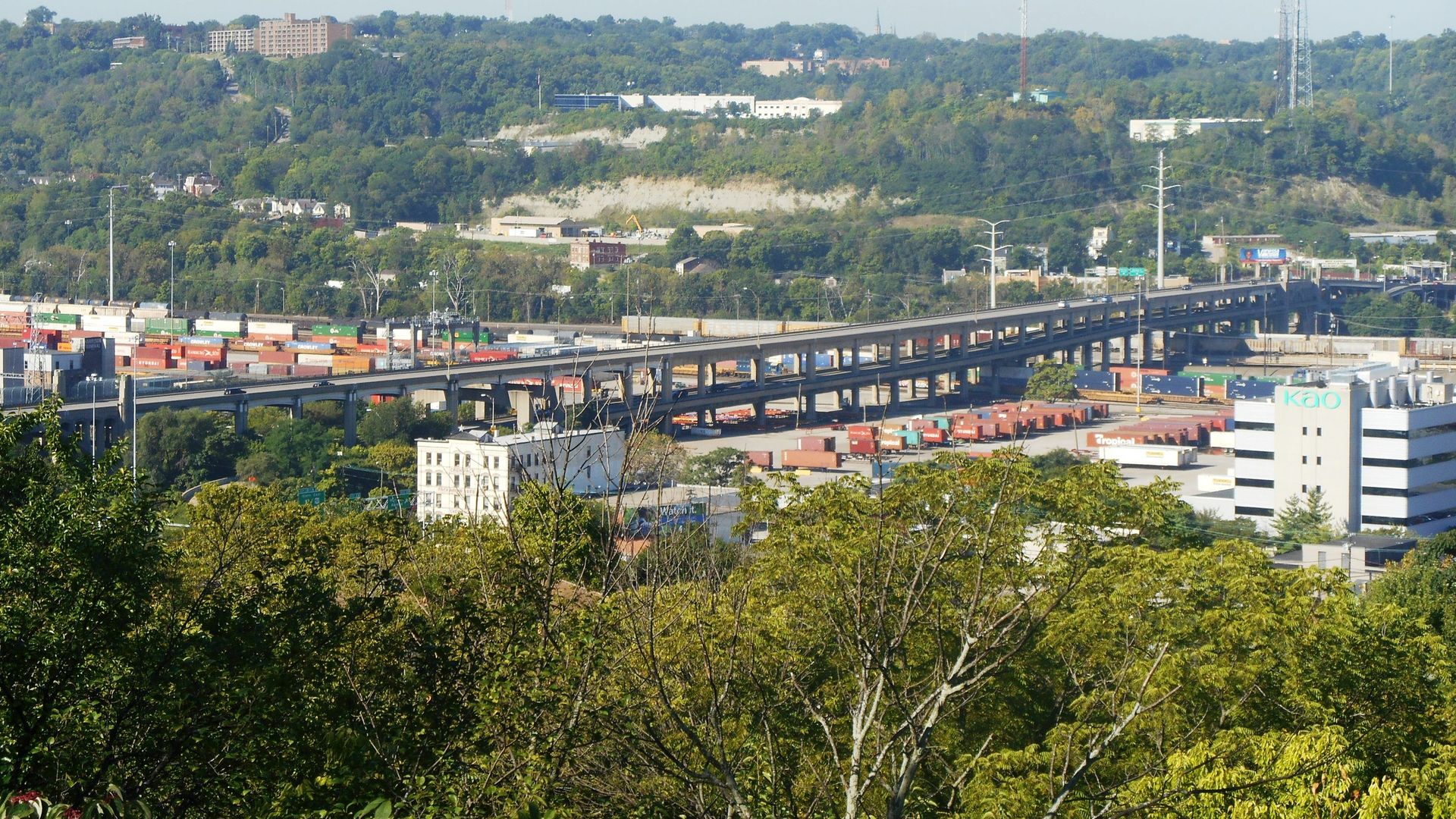 Wide view of a hilly, tree-filled landscape with an elevated highway in the foreground and shipping containers along the bottom; distant buildings and radio towers rise above treetops under blue sky.