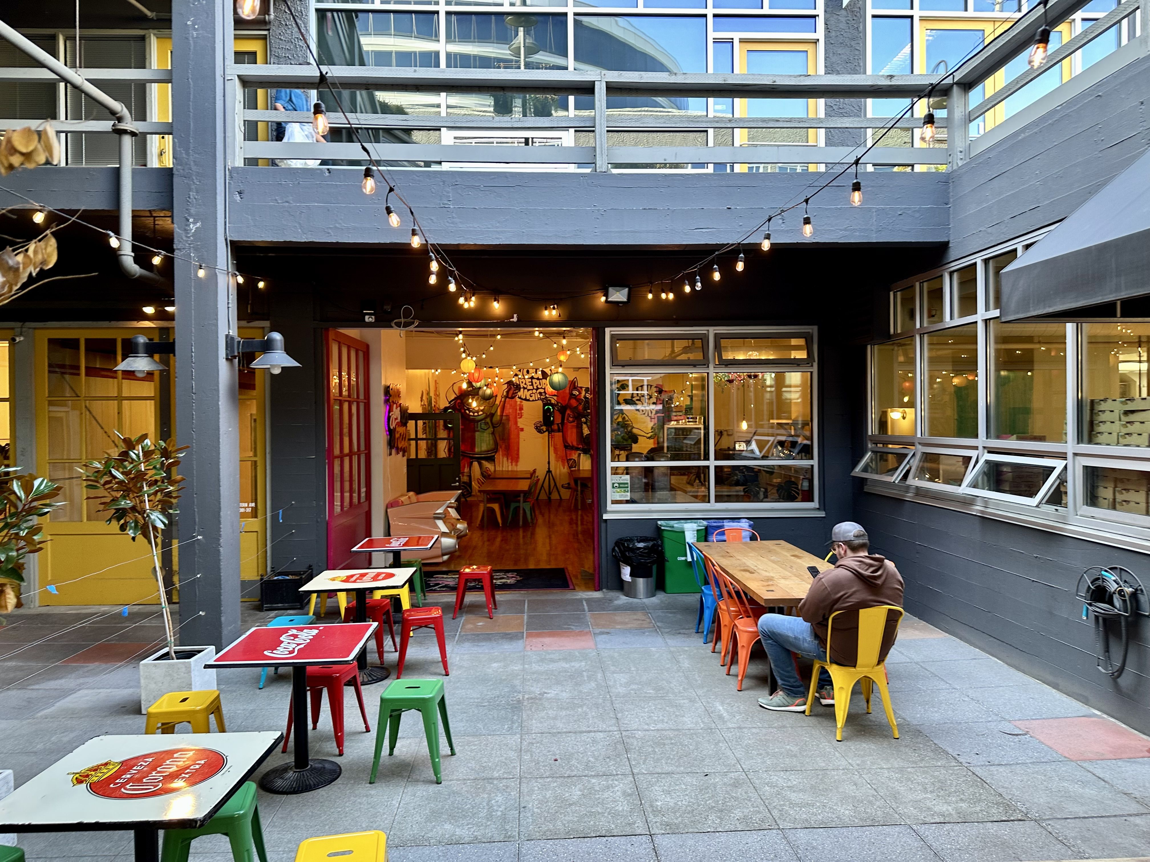 A courtyard with colorful tables and lights strung above, with an open storefront behind. 