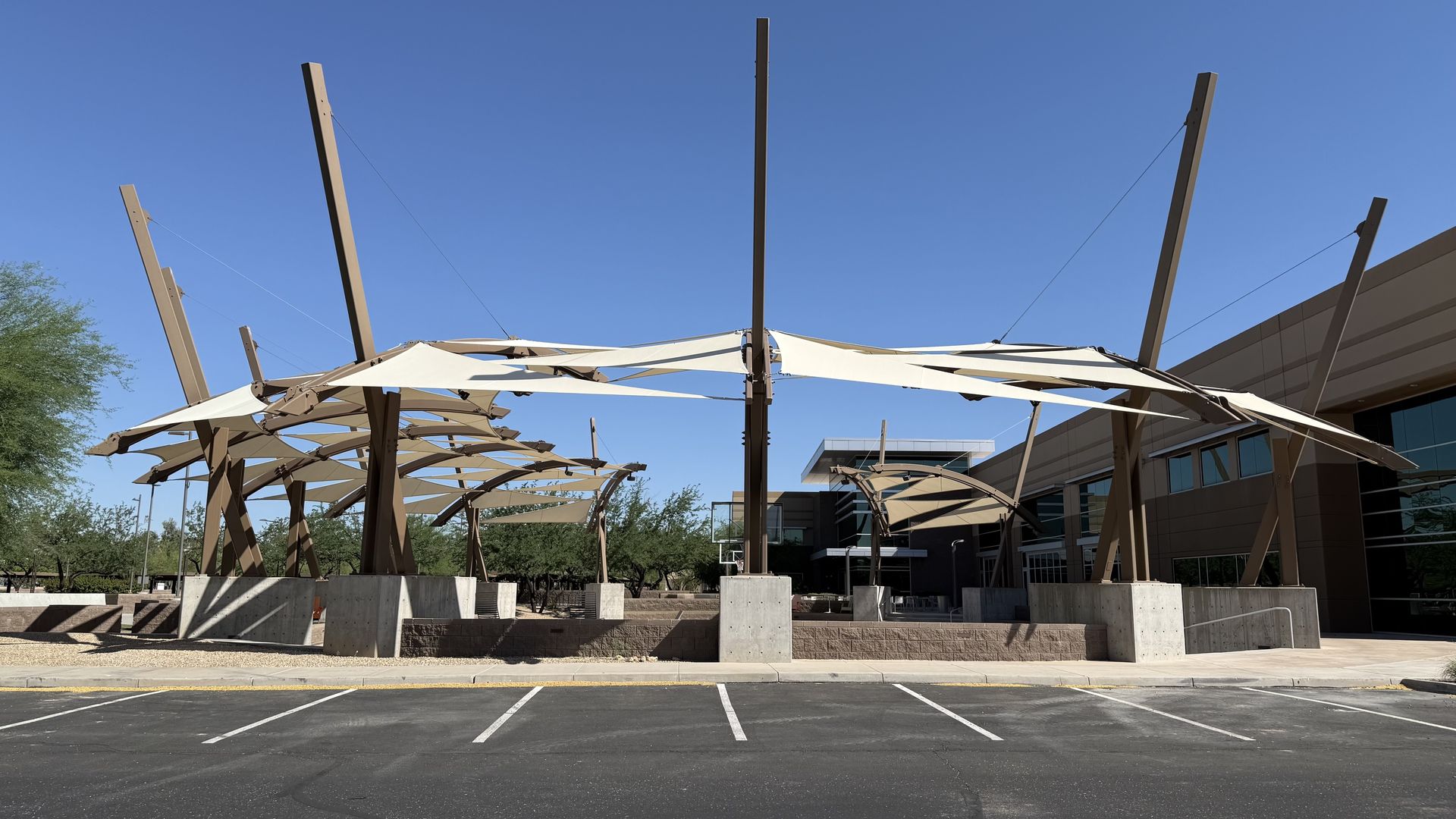 Sunlit outdoor plaza with tall brown metal beams and white fabric canopies stretched between them, adjacent to a modern building, over a paved parking lot under a clear blue sky.