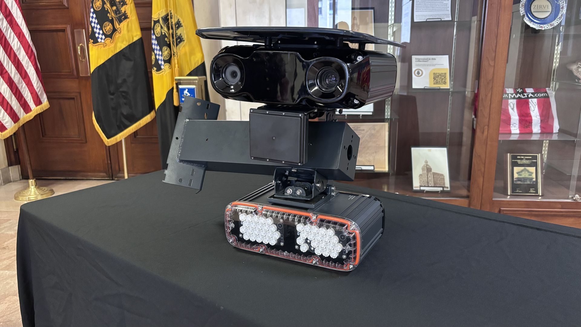 Glossy black security robot on a pedestal atop a black table, featuring dual camera lenses and a front LED array; flags and display cases visible in the background.