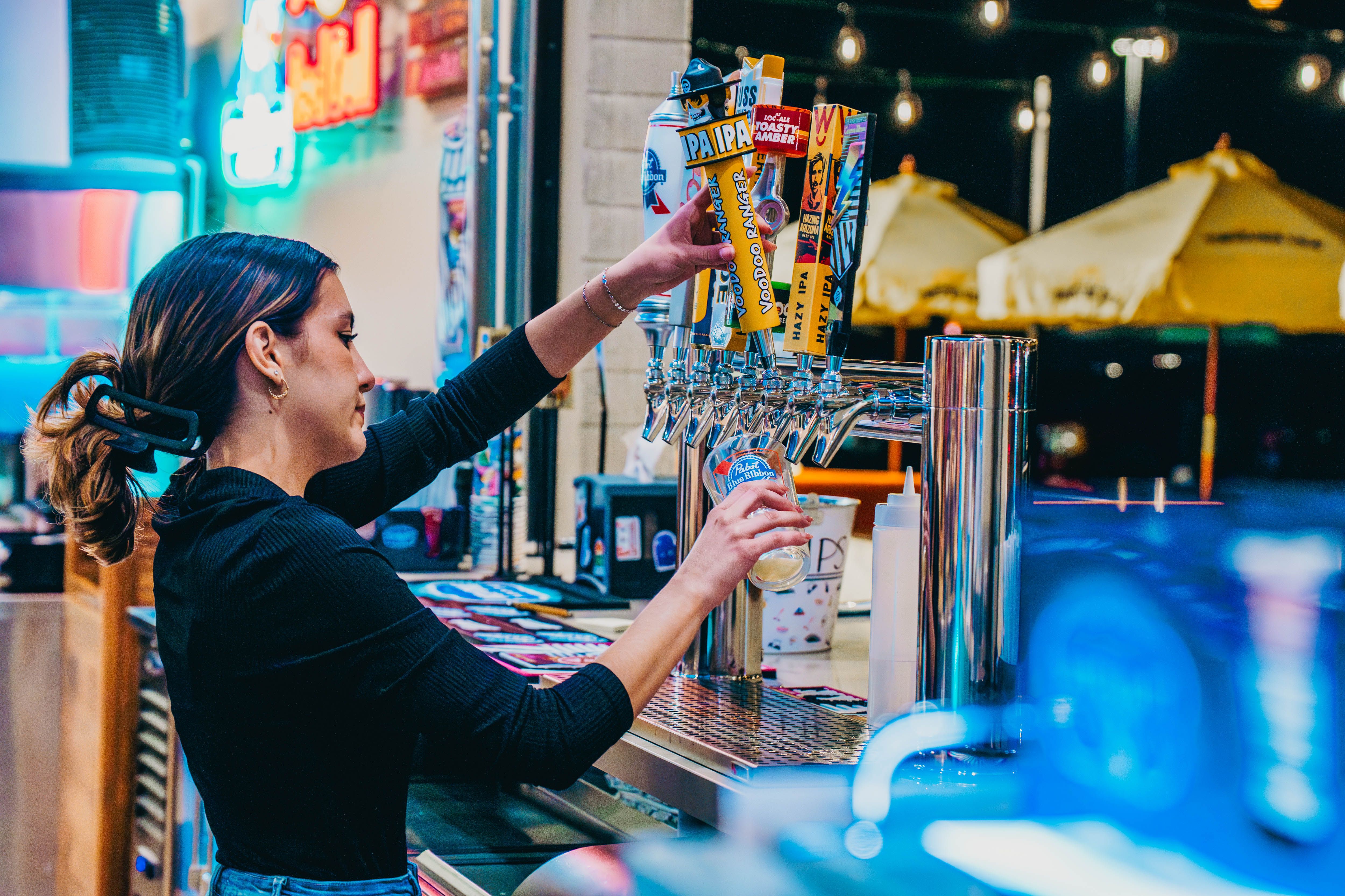Bar bartender pours beer from a row of colorful taps labeled IPA and Hazy IPA, with neon lights and outdoor umbrellas in the background.