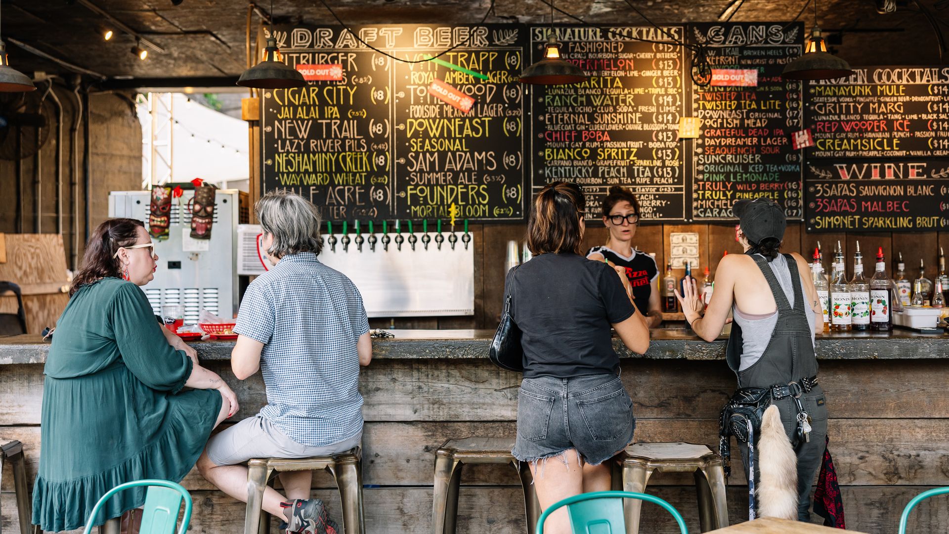 People sit at a rustic bar with chalkboard beer menus, warm hanging lamps, and dark wooden interior; a dog stands by the counter as customers order drinks.