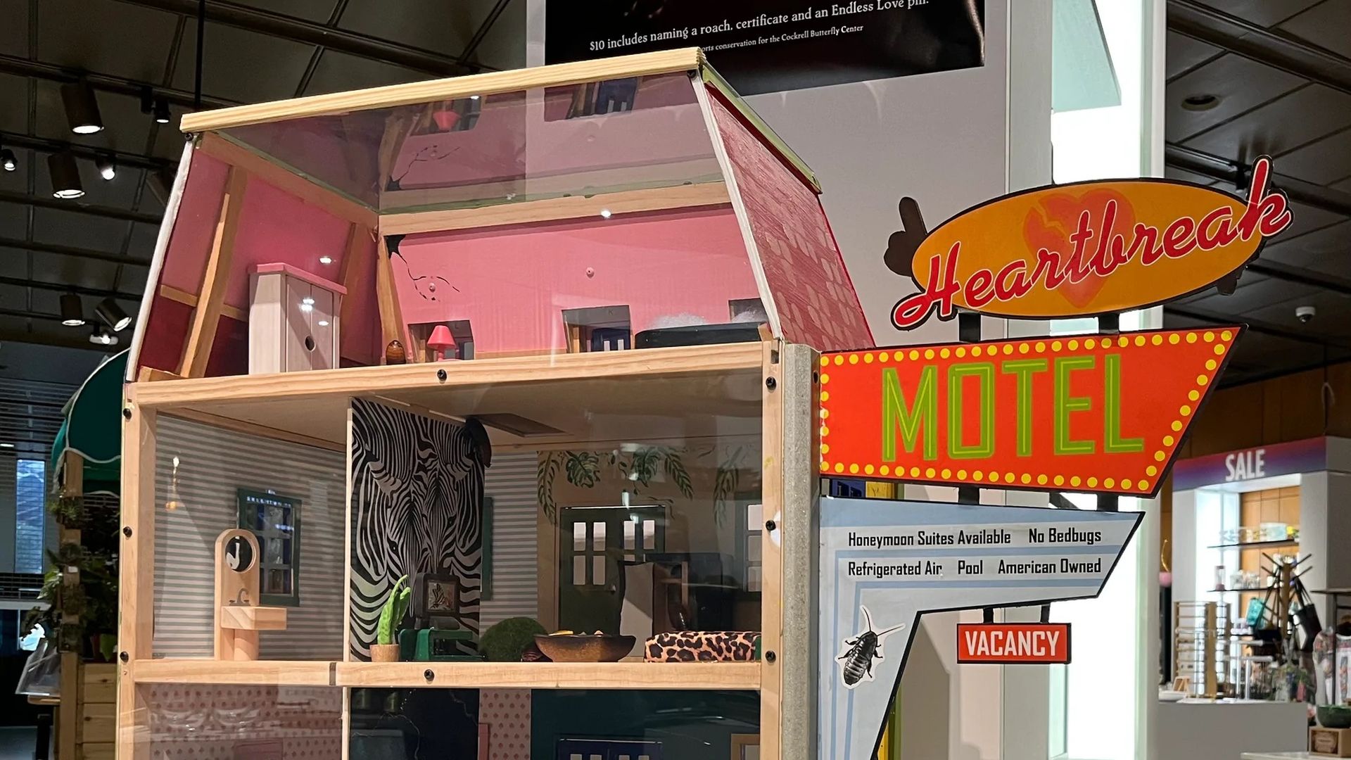 Model of a dollhouse with clear walls showing rooms with furniture, next to a retro neon style sign reading Heartbreak Motel with details like 'Honeymoon Suites Available' and 'Vacancy'.