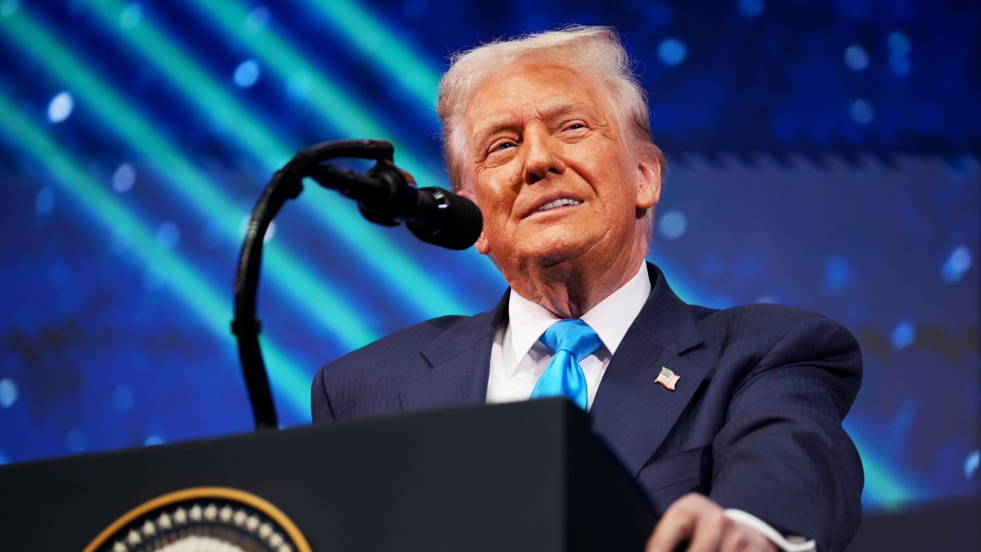 Trump grips the side of a podium as he speaks into a microphone while wearing a dark suit, a light blue tie, a white collared shirt and an American flag pin. 