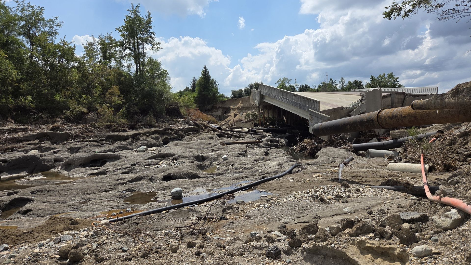 Partially collapsed concrete bridge over a rocky, muddy creek bed with exposed rusted pipes and scattered debris; dense trees line the left and a blue sky with clouds above.