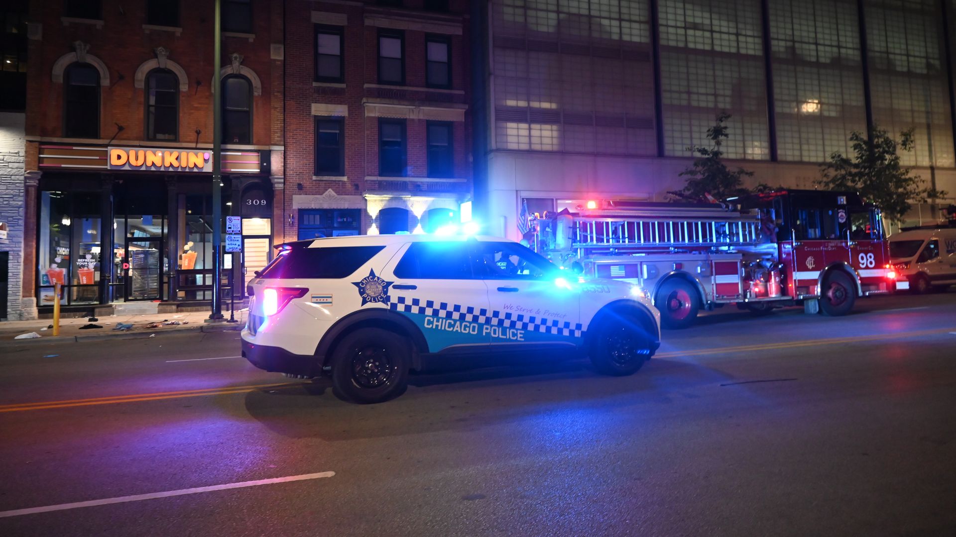 Photo of a police car with its lights on in front of a restaurant and a fire truck. 