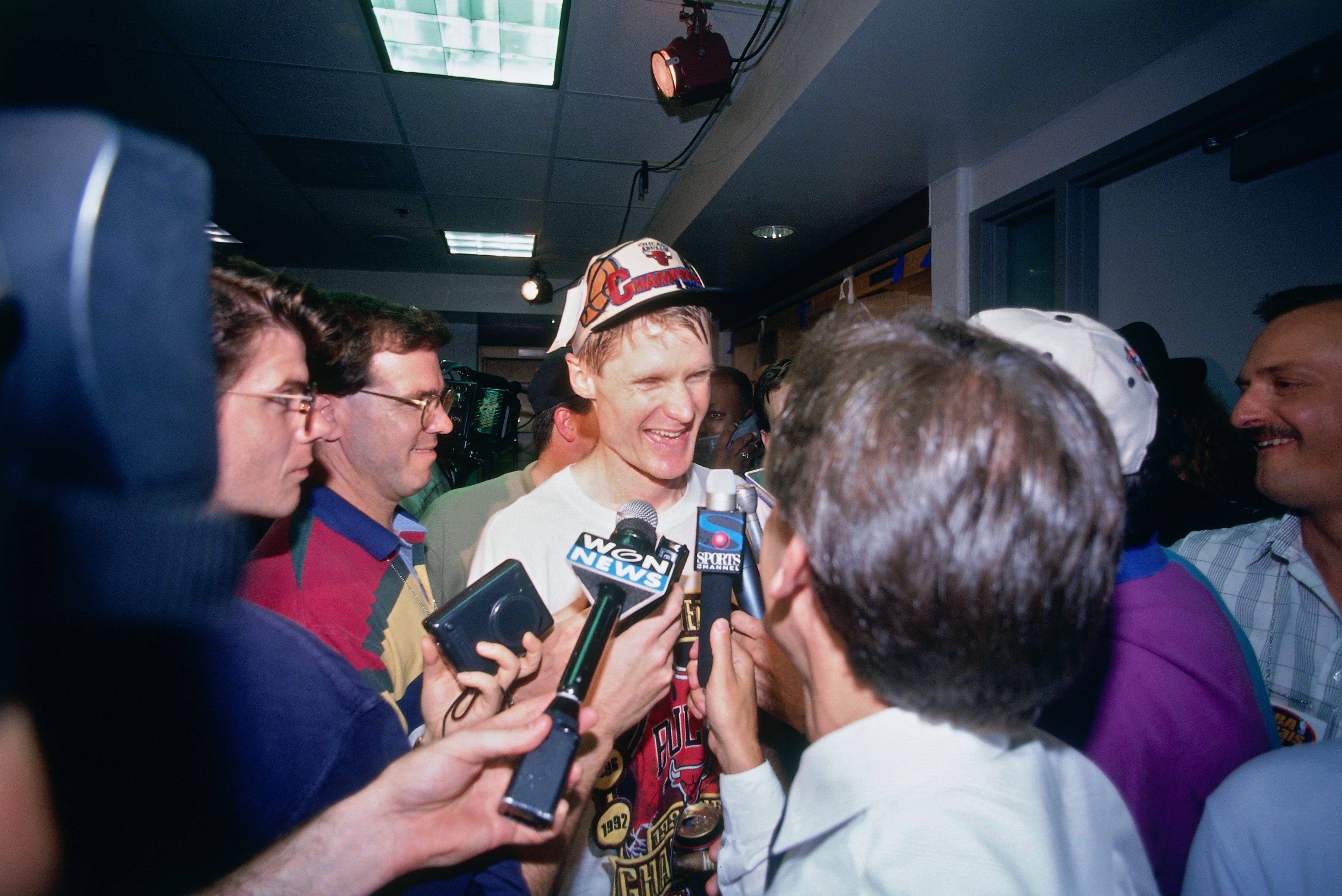 A smiling man wearing a white Chicago Bulls cap is surrounded by reporters and microphones in a crowded indoor hallway, with ceiling lights and cameras capturing the scene.