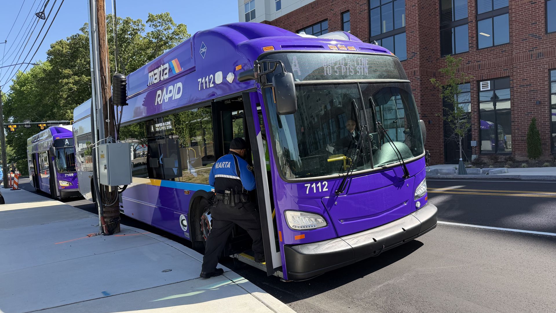 Two purple MARTA Rapid buses on a street; foreground bus #7112 doors open as a driver boards, second bus #7109 behind. A brick building, trees, and power lines.