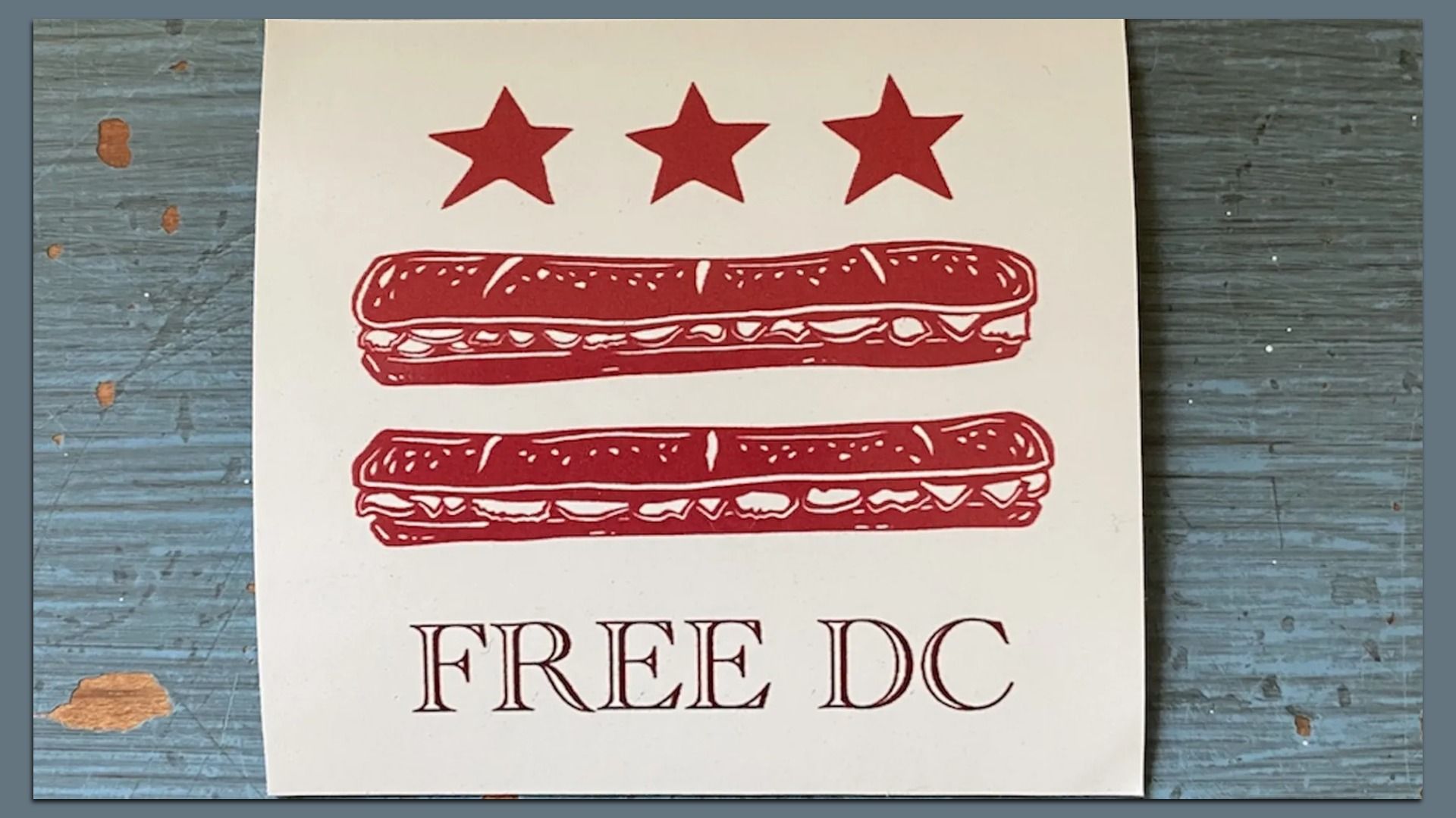 Illustration of three red stars above two red sandwiches on white paper, with the text "FREE DC" below, placed on a worn blue wooden surface.