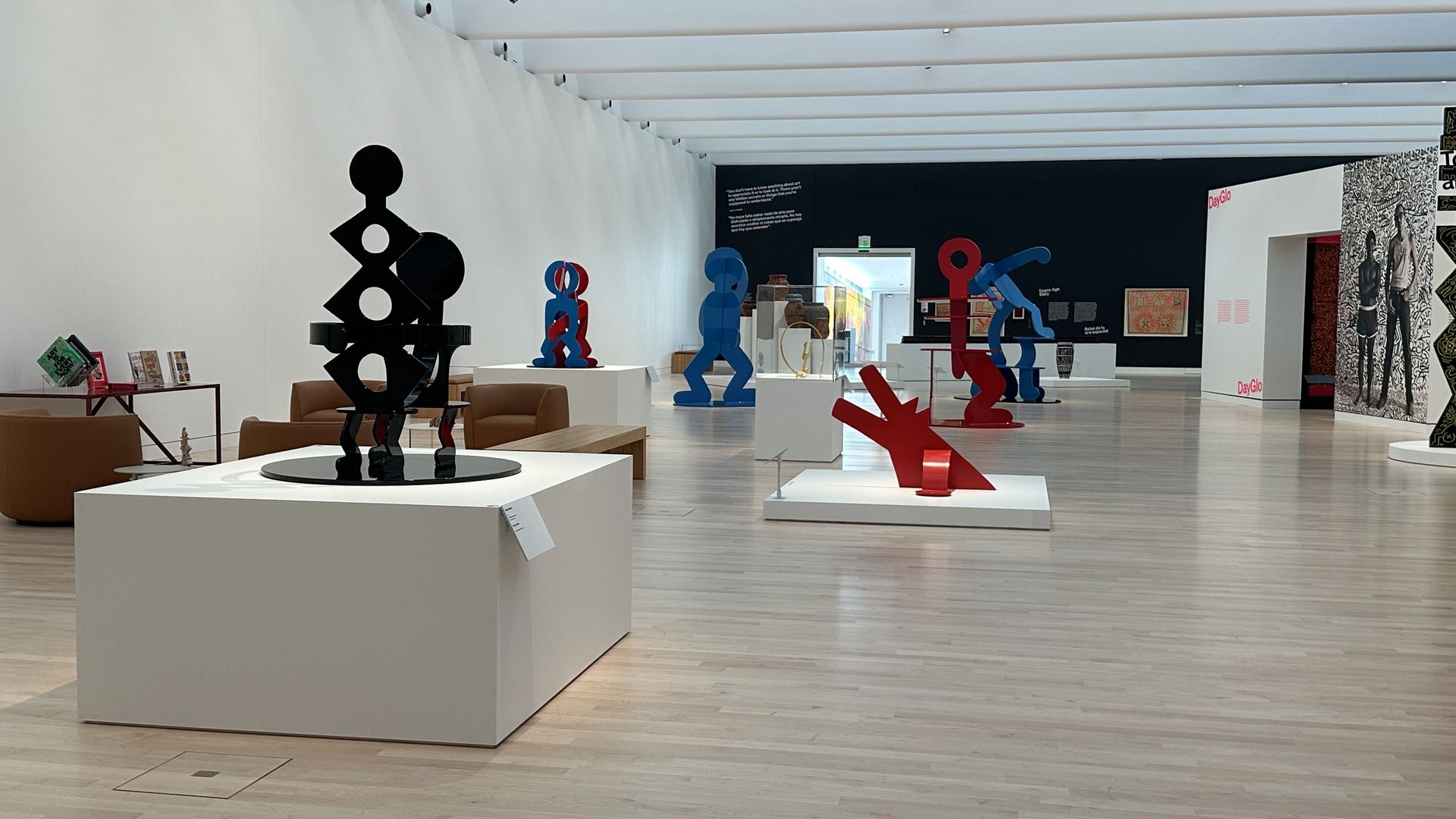 Bright art gallery with white walls and light wood floor, featuring abstract black, blue, and red sculptures on white pedestals; a dark wall with text panels in the background.