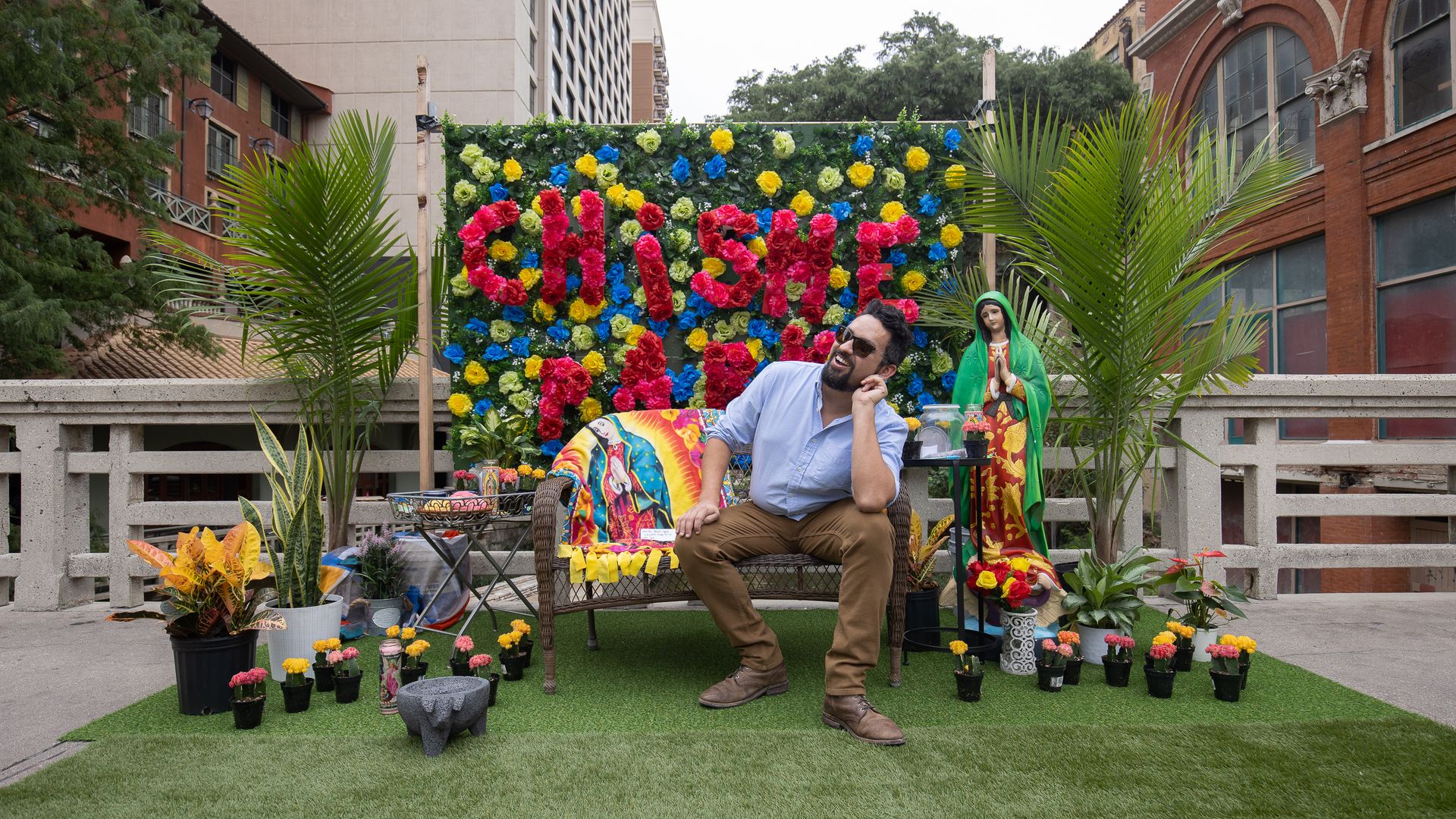 A man sits at a mini park set up with Latino motifs like a background that says "chisme."