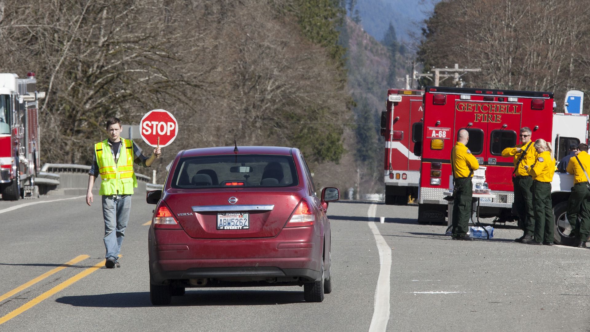 Man in yellow safety vest holding a stop sign on a road with a red car stopped. Firefighters in yellow jackets stand near two red Getchell Fire trucks on the roadside.