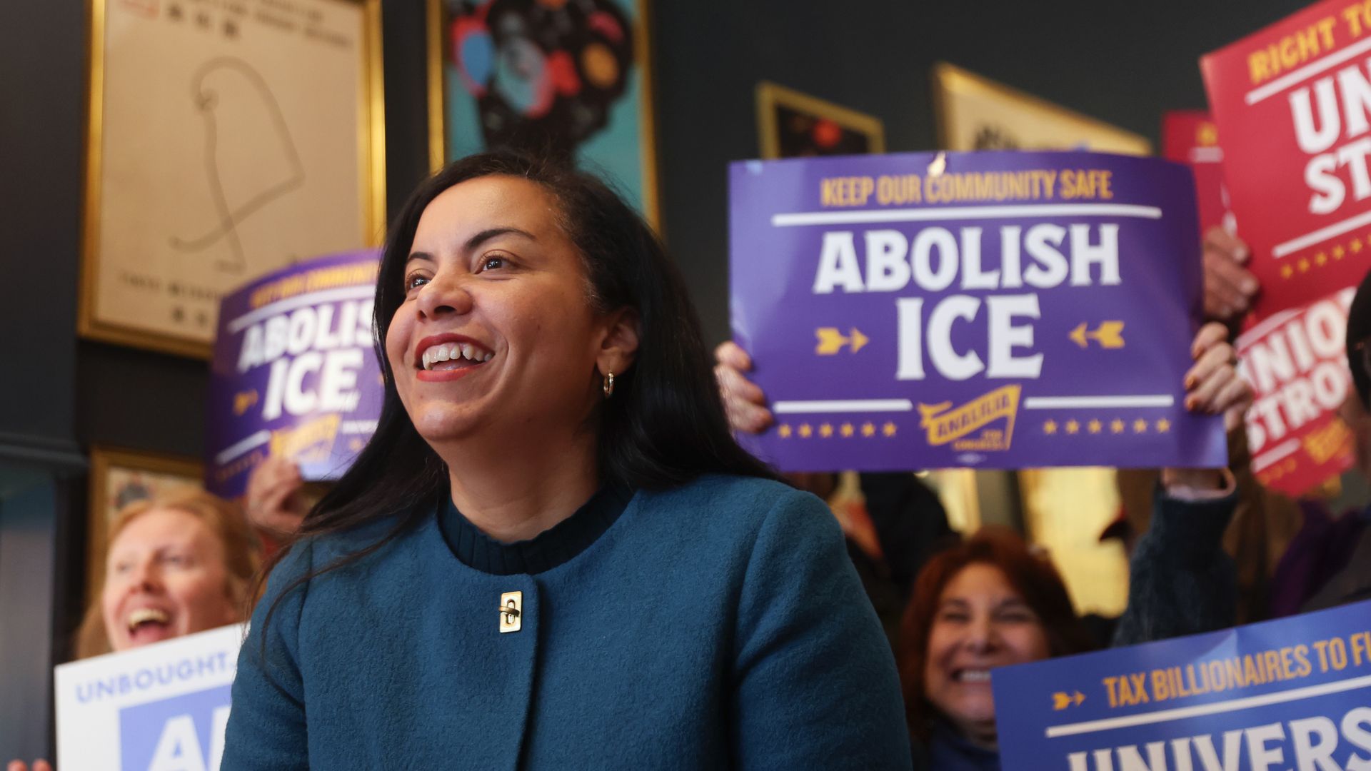 Smiling woman in a teal coat at an indoor rally with people holding purple signs reading "ABOLISH ICE" and other signs advocating community safety and university funding.